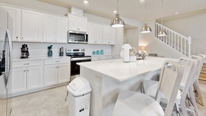 Una cucina moderna con mobili bianchi, un'isola centrale, elettrodomestici in acciaio inossidabile e illuminazione a sospensione.
