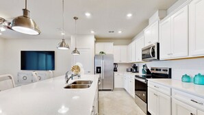 Una cucina moderna con mobili bianchi, elettrodomestici in acciaio inossidabile, un'isola centrale, illuminazione a sospensione e una televisione a schermo piatto montata a parete.