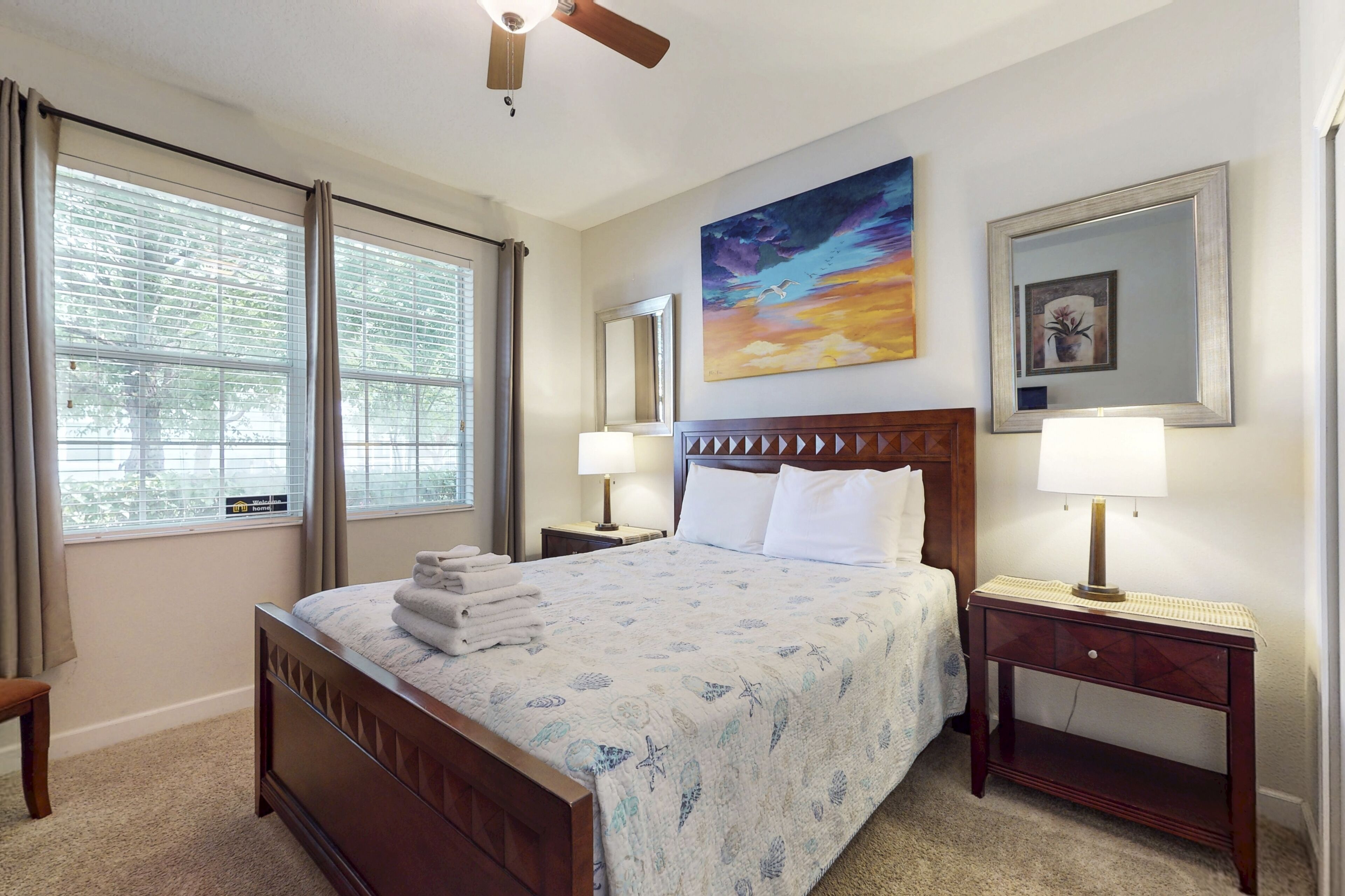 A hotel room with a large bed, two bedside tables with lamps, a ceiling fan, a large window with curtains, and a painting on the wall.