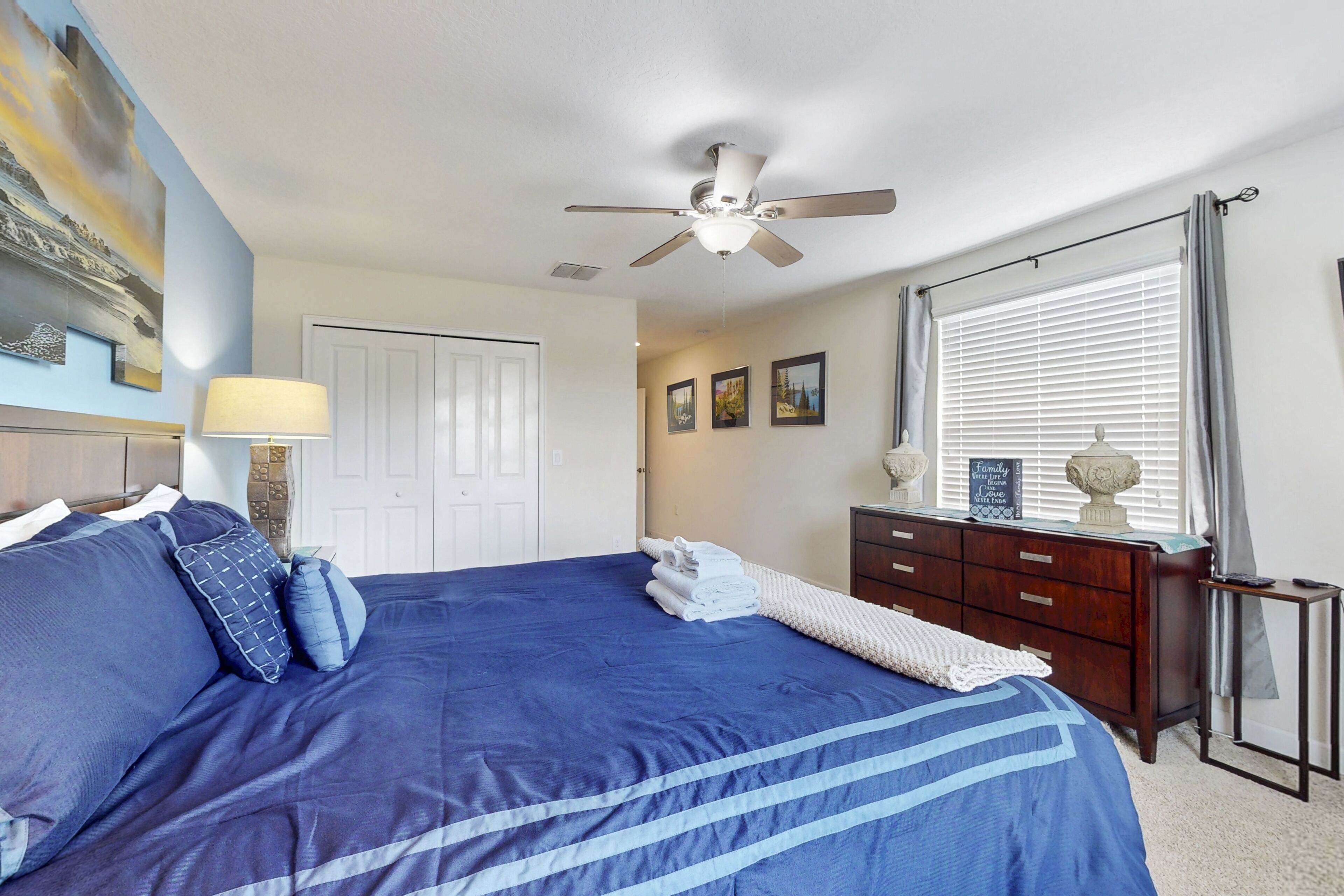 A bedroom with a large bed, a ceiling fan, a dresser, and a window with curtains.