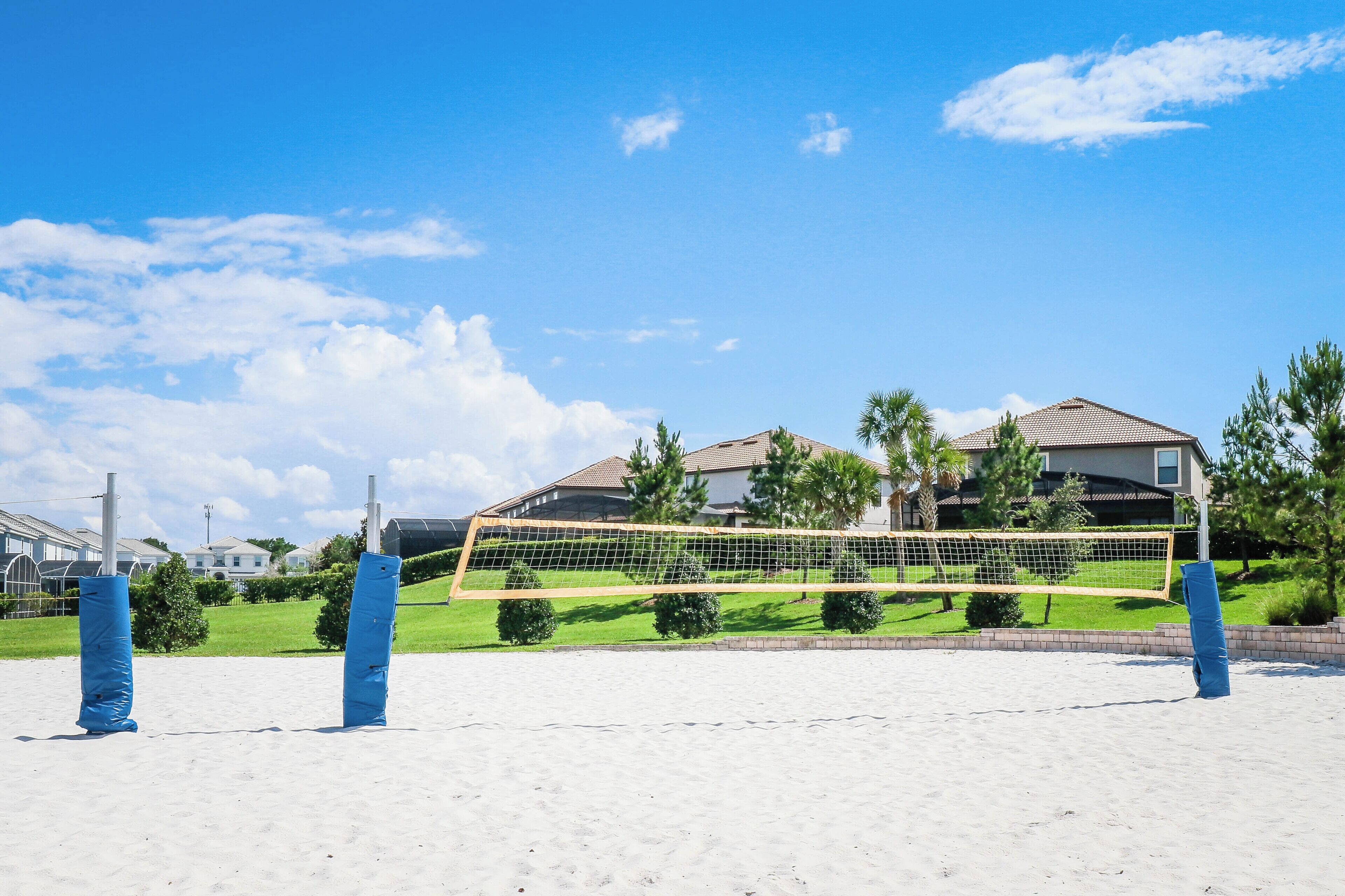 Una cancha de voleibol de playa con una red, rodeada de arena y palmeras, con casas al fondo.