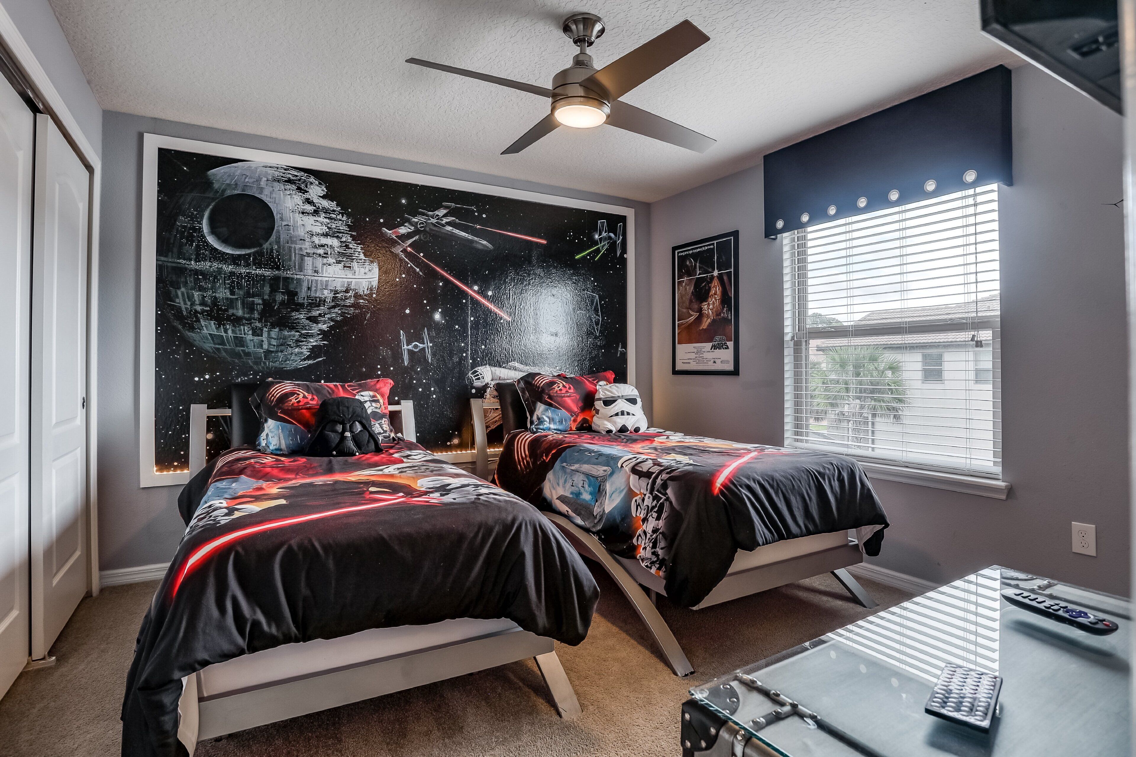 A bedroom with two beds, a ceiling fan, a window with blinds, and a desk with a remote control.