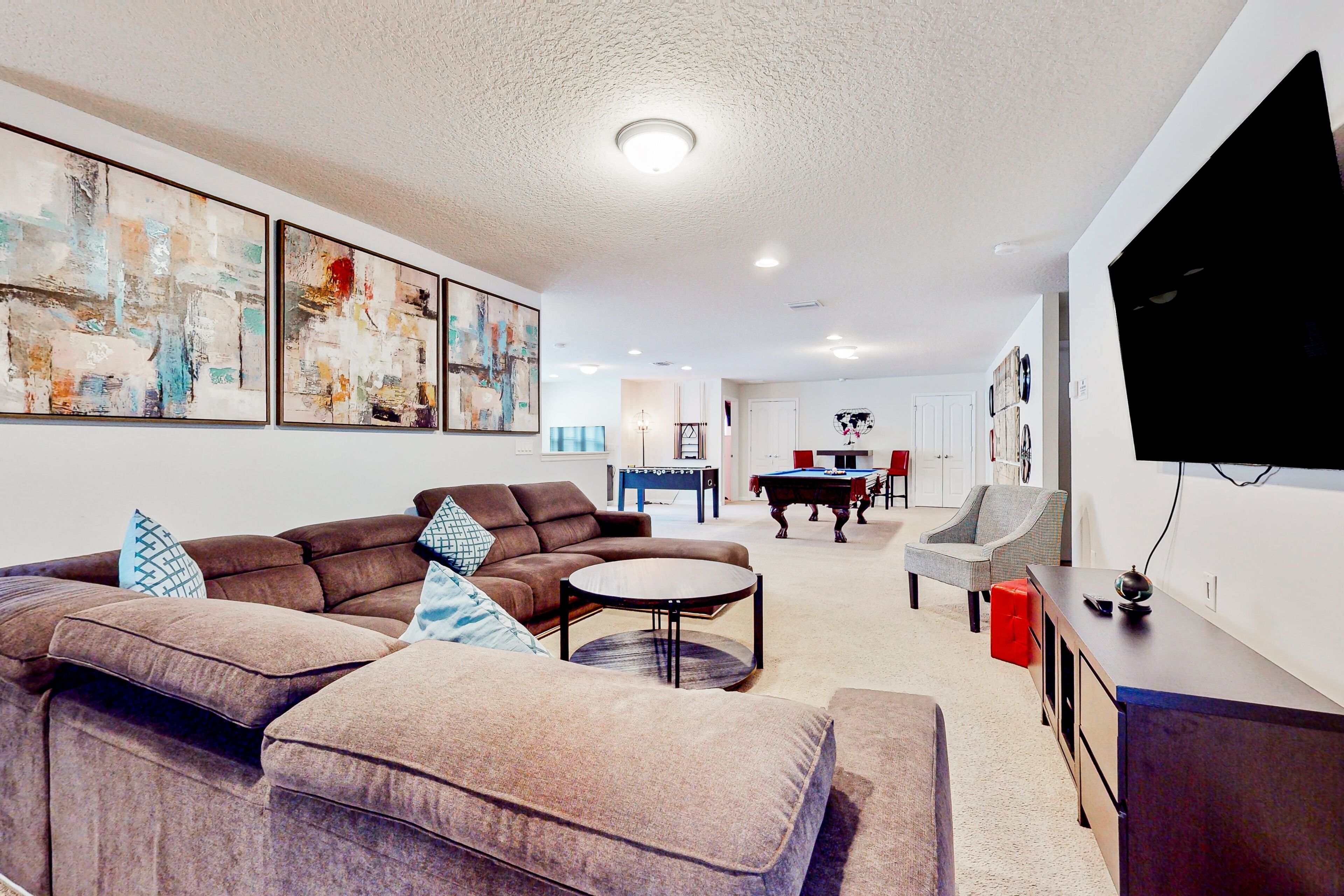A modern living room with a sectional sofa, a round coffee table, and a flat-screen TV. There is a pool table and a dining area in the background.