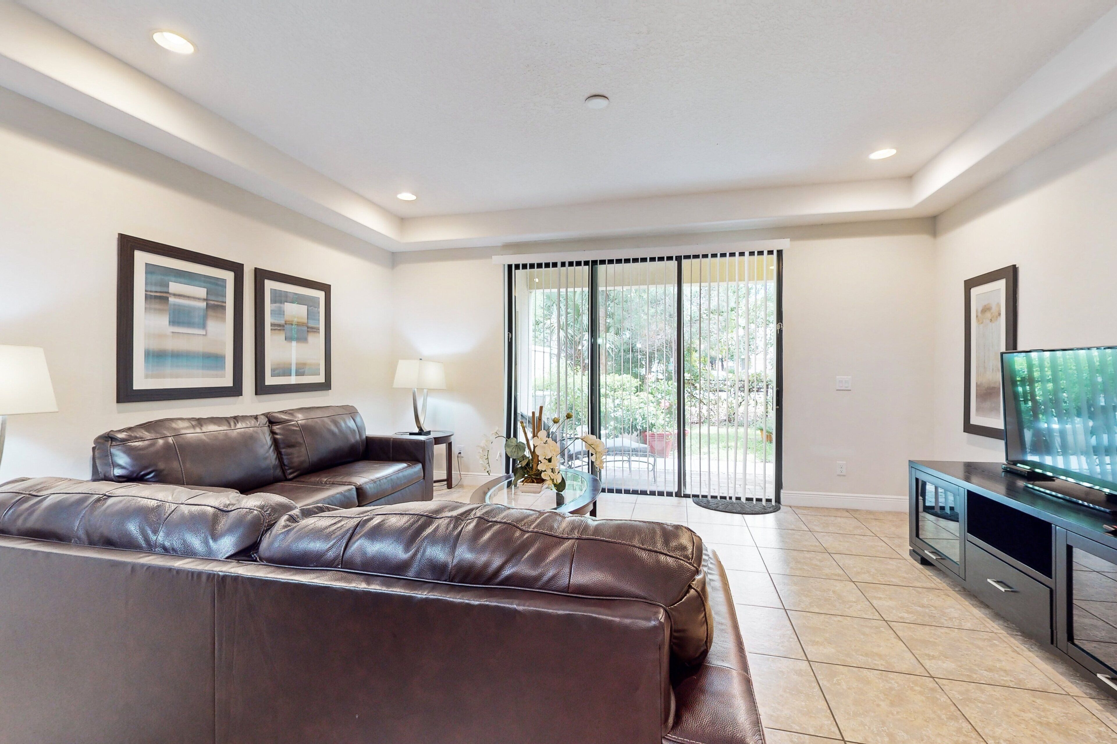 A living room with a brown leather sofa, a glass coffee table, a TV on a stand, and a sliding glass door leading to a patio.