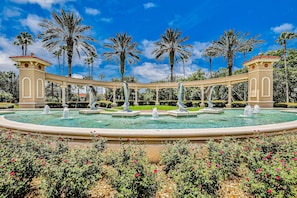 Ein runder Brunnen mit Statuen, umgeben von Palmen und wolkenlosem Himmel.