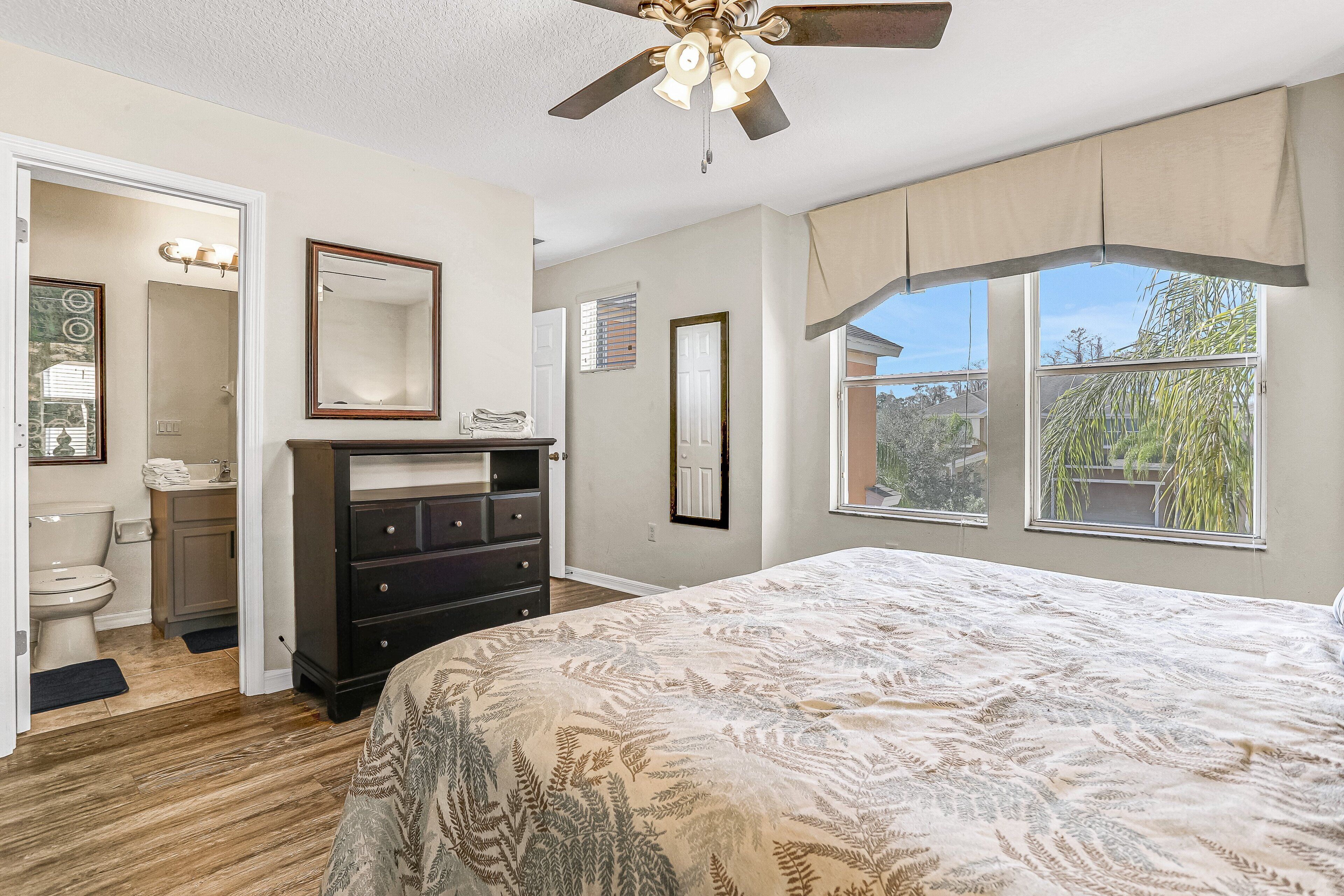 A bedroom with a bed, dresser, ceiling fan, and large windows.