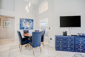 A dining area with a round table, blue upholstered chairs, a large blue artwork, a TV on a blue cabinet, and a window with blinds. - Beautiful Home With Private Pool, Screened Lanai, Wifi, Private W/d, and AC (Kissimmee)