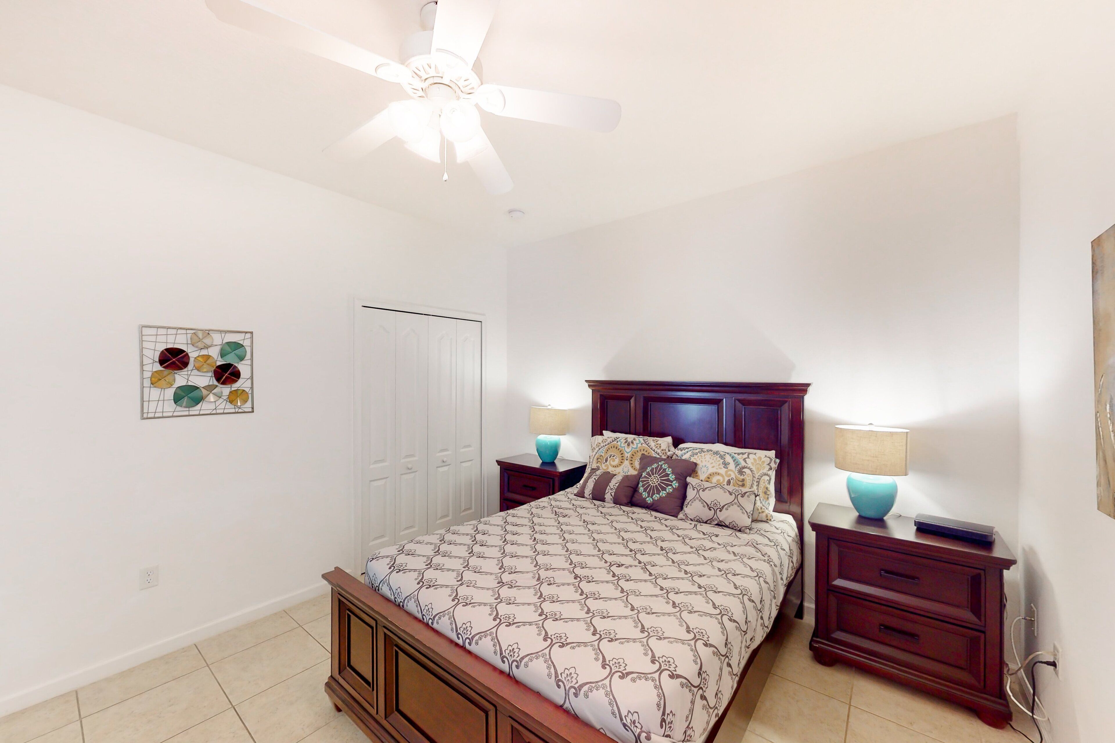 A bedroom with a bed, nightstands, a ceiling fan, a door, and a wall art piece.