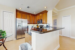 A kitchen with wooden cabinets, a stainless steel refrigerator, a microwave, and a granite countertop island. - Top-floor 2BR Condo With Golf Course Views, Garage & Reunion Resort Amenities (Kissimmee)