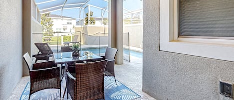A covered patio area with a glass table and wicker chairs, overlooking a swimming pool and residential buildings.