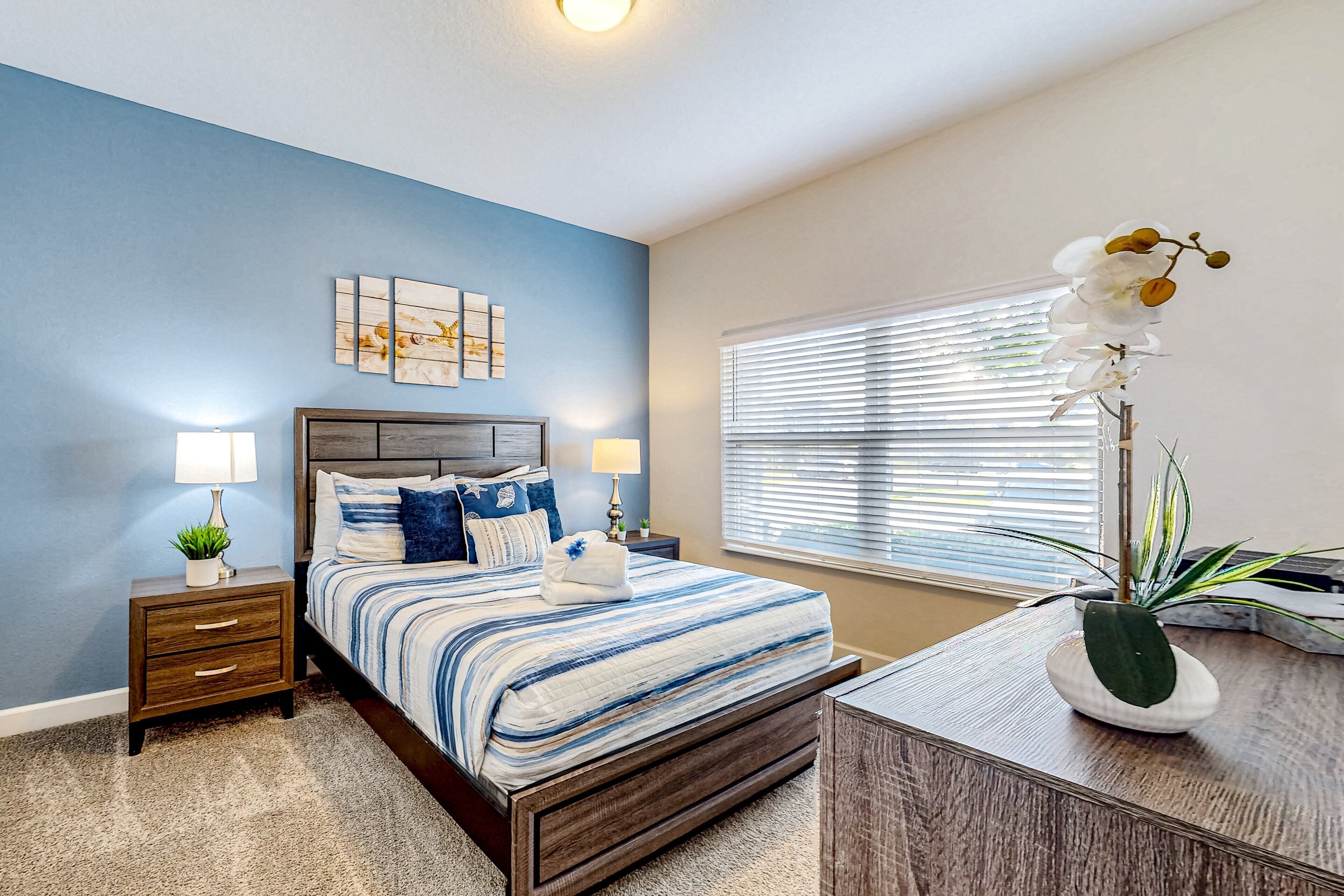 A bedroom with a bed, nightstands, a desk, a window with blinds, and a wall with a painting.