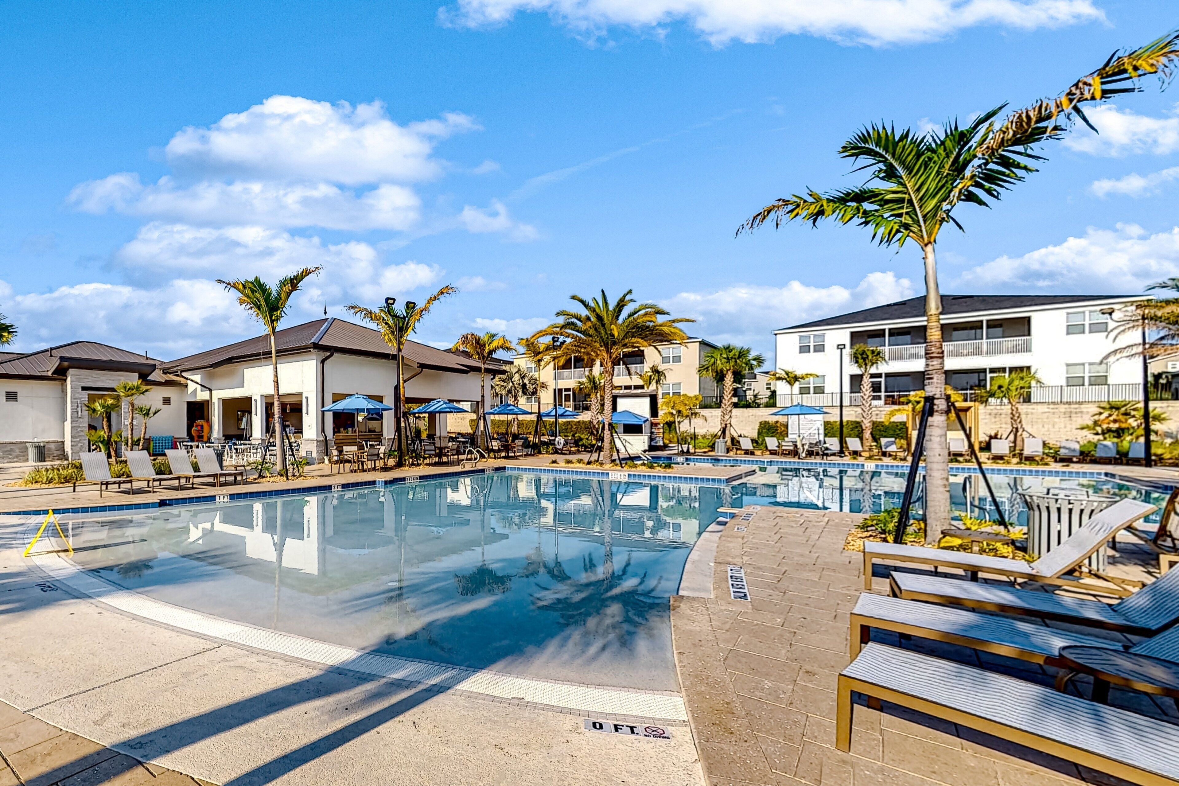 A swimming pool with lounge chairs and umbrellas, surrounded by palm trees and buildings.