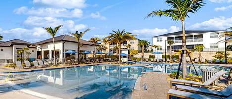 A swimming pool with lounge chairs and umbrellas, surrounded by palm trees and buildings.