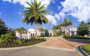 A gated entrance with palm trees, a sign for 'The Enclaves Amenity Center,' and a flagpole with an American flag. - Family-friendly Home With Private Pool, Wifi, Central AC, & Private Washer/dryer (Davenport)
