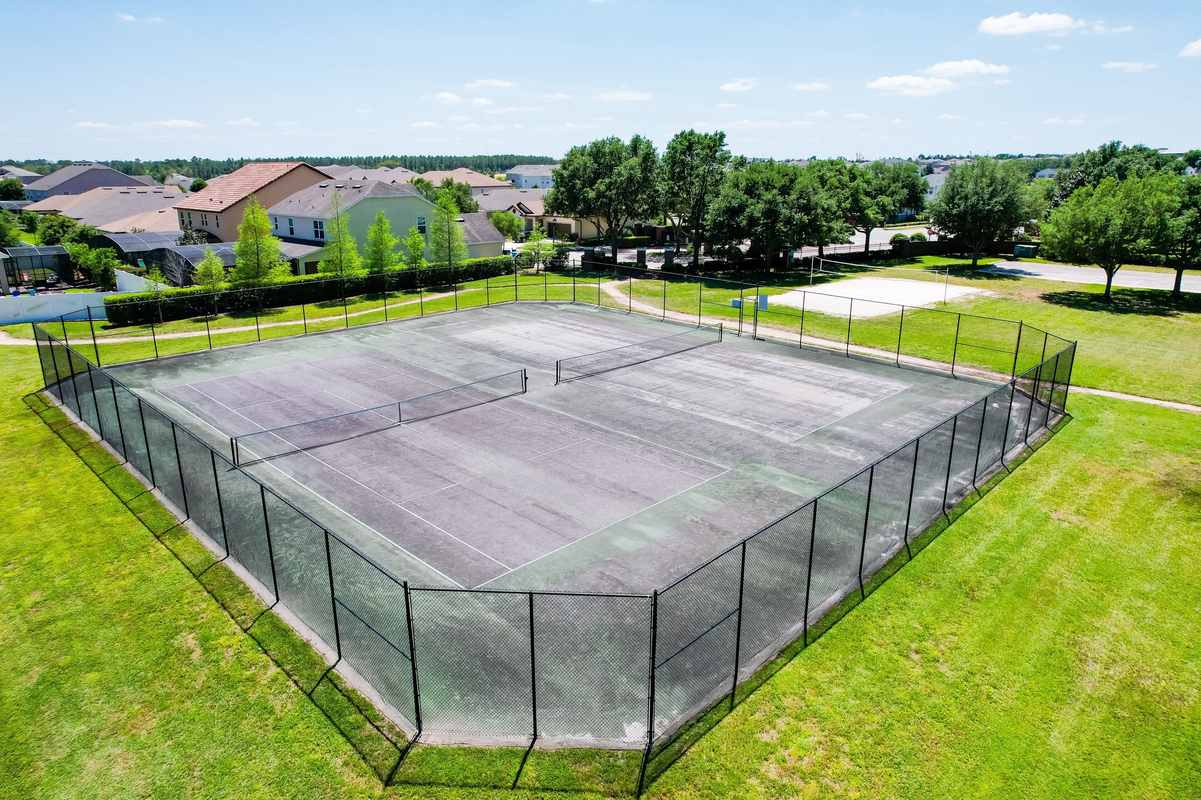 Una cancha de tenis rodeada por una valla, con casas y árboles al fondo.