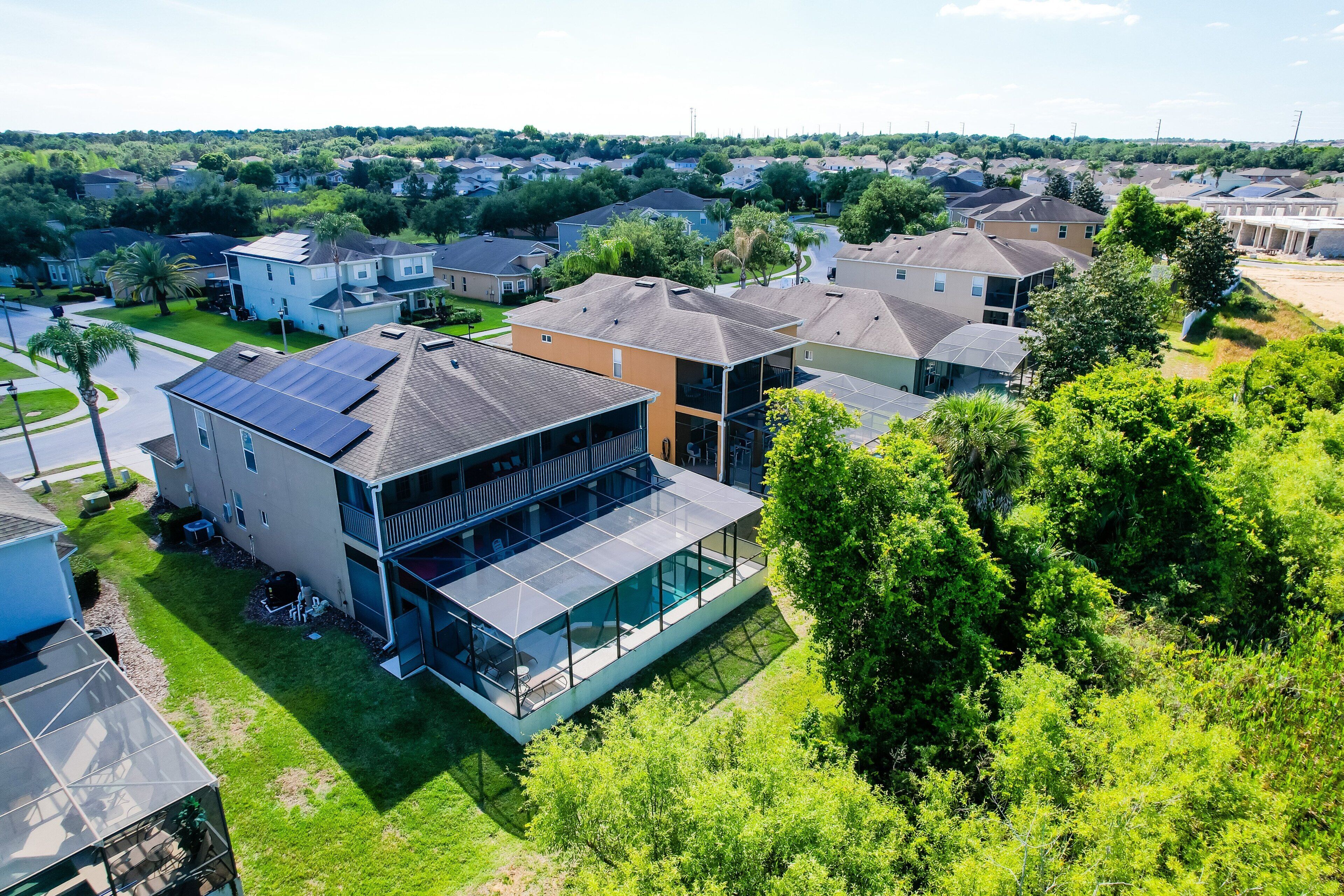 A modern residential building with a glass conservatory, surrounded by lush greenery and other houses.
