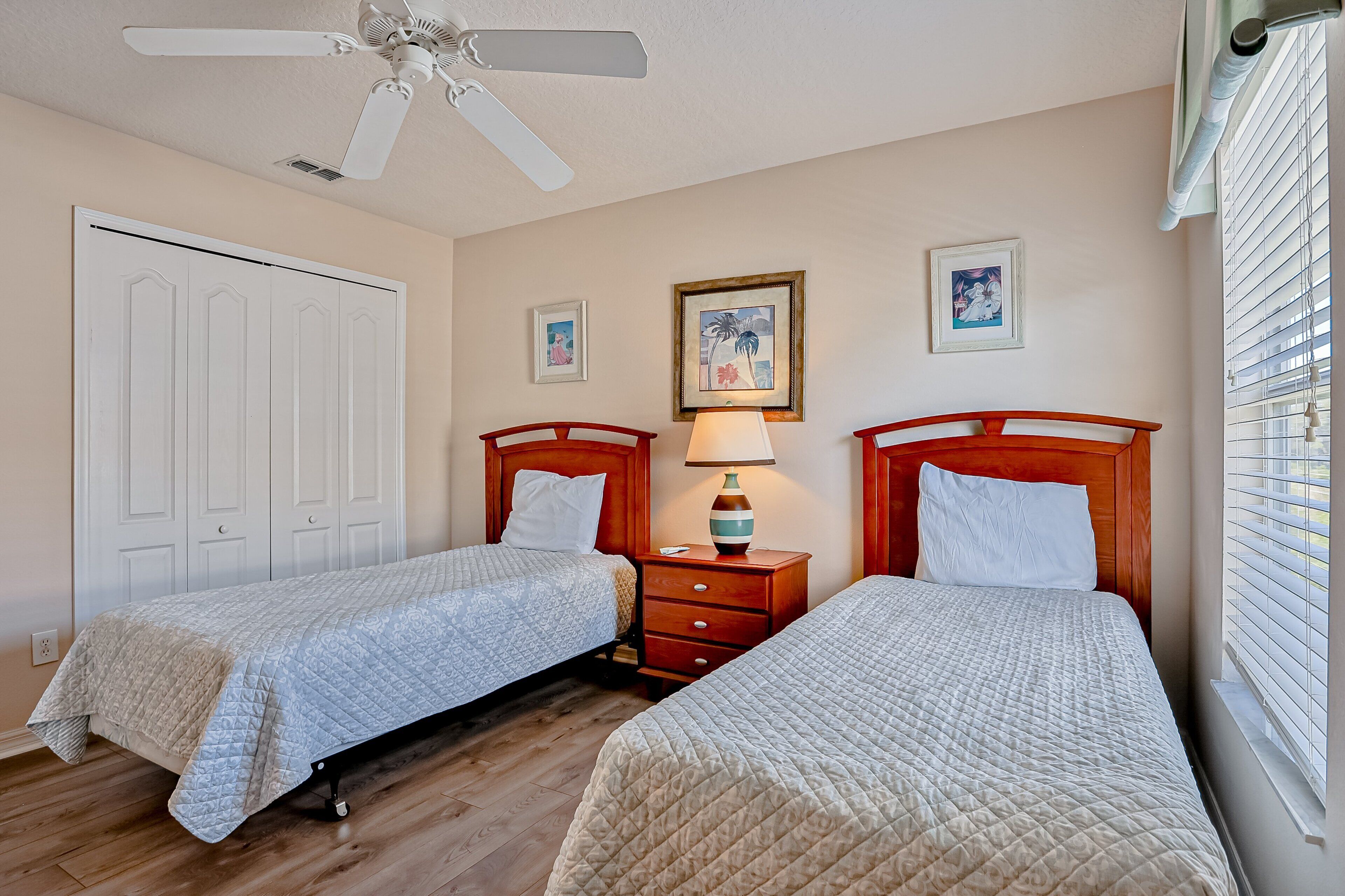 A bedroom with two beds, a ceiling fan, a nightstand with a lamp, and a window with blinds.