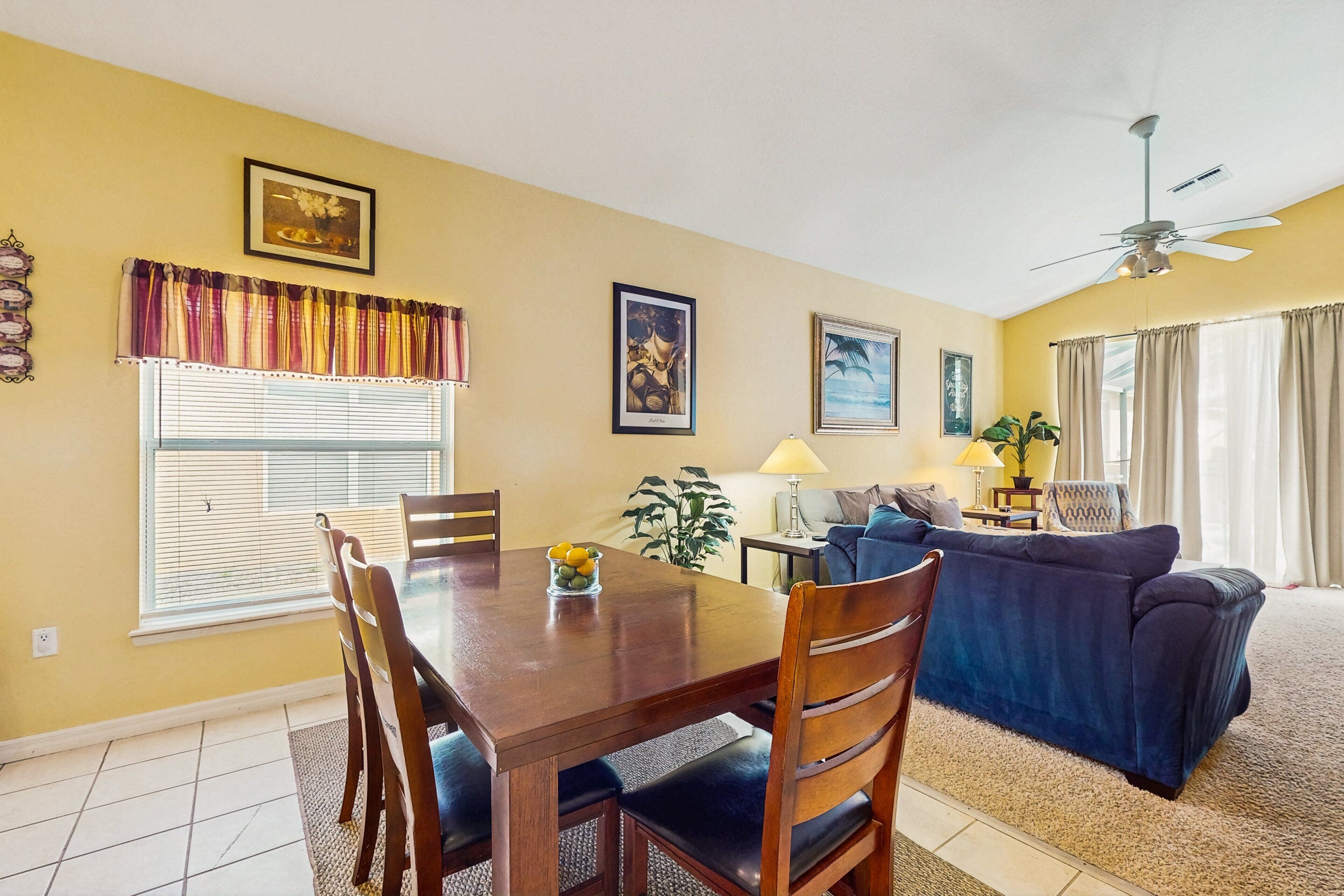 A dining area with a table, chairs, and a vase of flowers. There is a sofa and a lamp in the living area.