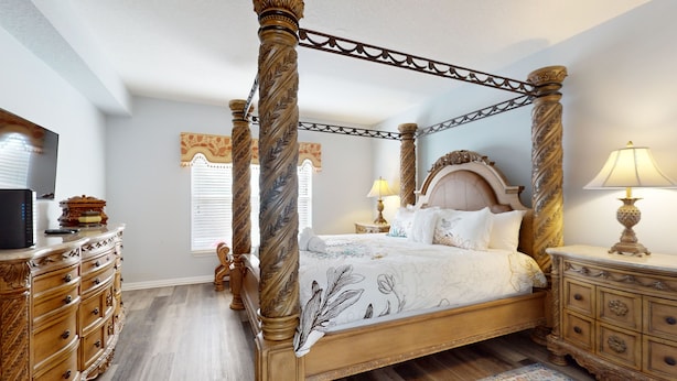 A four-poster bed with a floral bedspread, a wooden dresser with drawers, a nightstand with a lamp, and a television on a dresser. - Wonderful Home For Theme Park Fun W/private Pool, Game Room, Wifi, AC, Patio, WD (Davenport)
