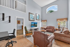 A spacious living room with a high ceiling, a staircase, and a large window providing natural light. - Wonderful Home For Theme Park Fun W/private Pool, Game Room, Wifi, AC, Patio, WD (Davenport)