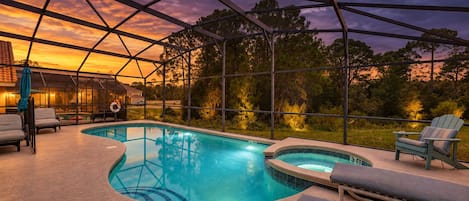 A swimming pool with a hot tub, surrounded by lounge chairs and a covered area with a view of trees and a sunset.