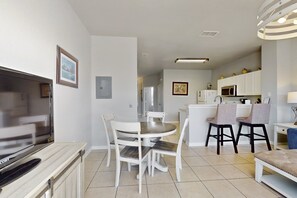 Een moderne woonkamer met een flatscreen televisie, een eettafel met stoelen, een keuken met een magnetron, en een kleine zithoek met een bankje.