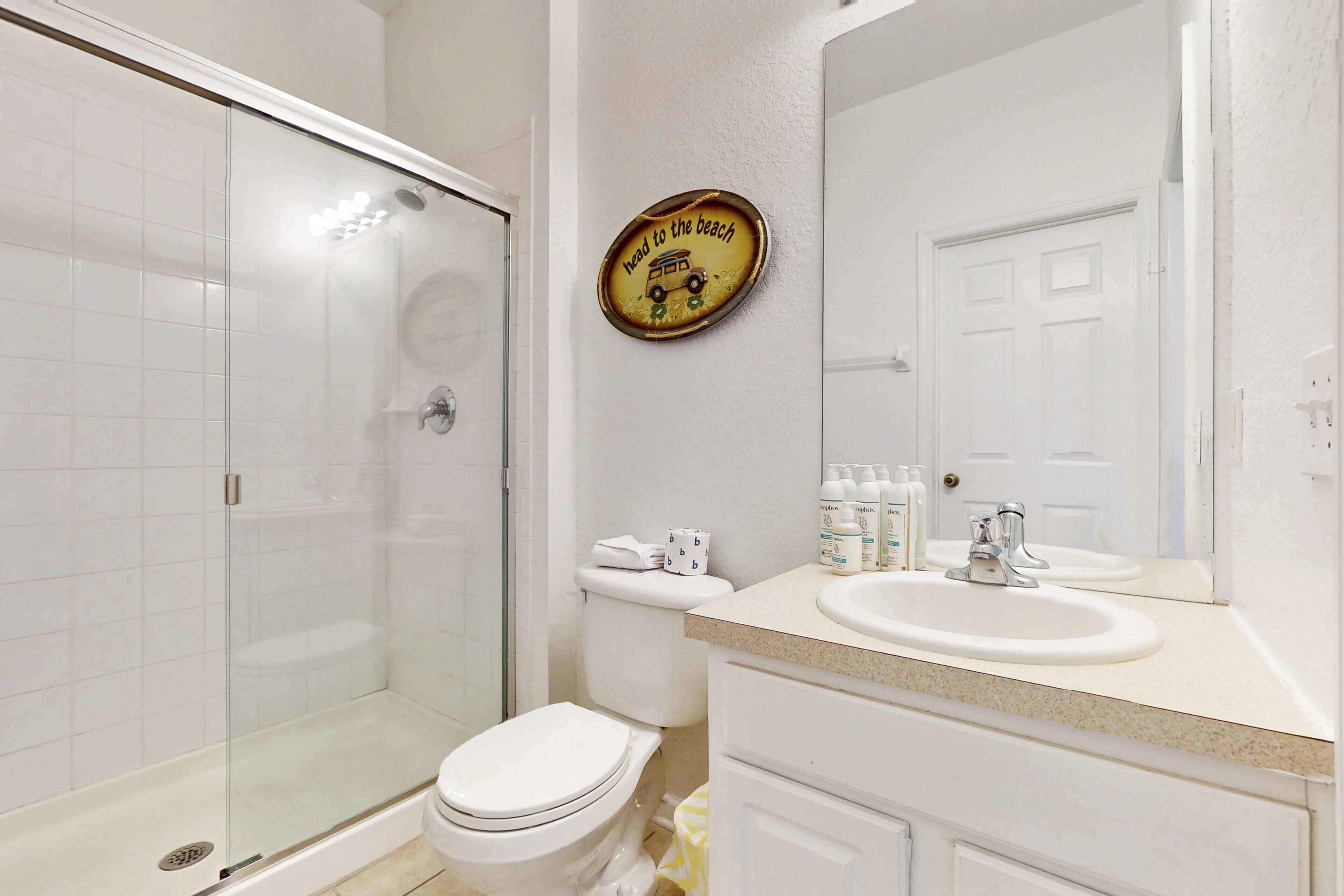 A bathroom with a glass shower enclosure, a toilet, a sink with a mirror, and a wall-mounted sign.