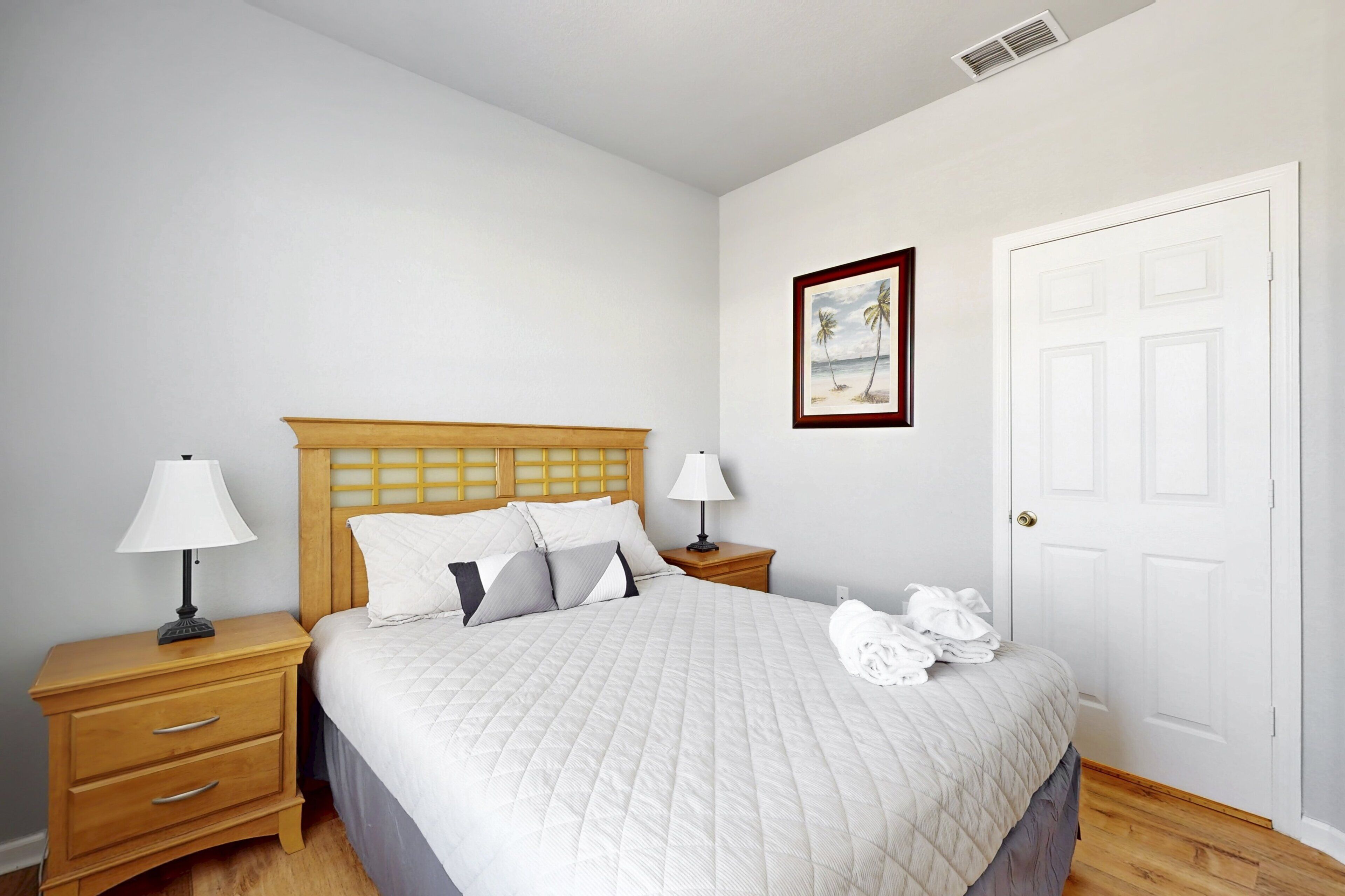 A neatly made bed with a quilted blanket, two bedside lamps, a wooden nightstand with drawers, a framed picture on the wall, and a white door.