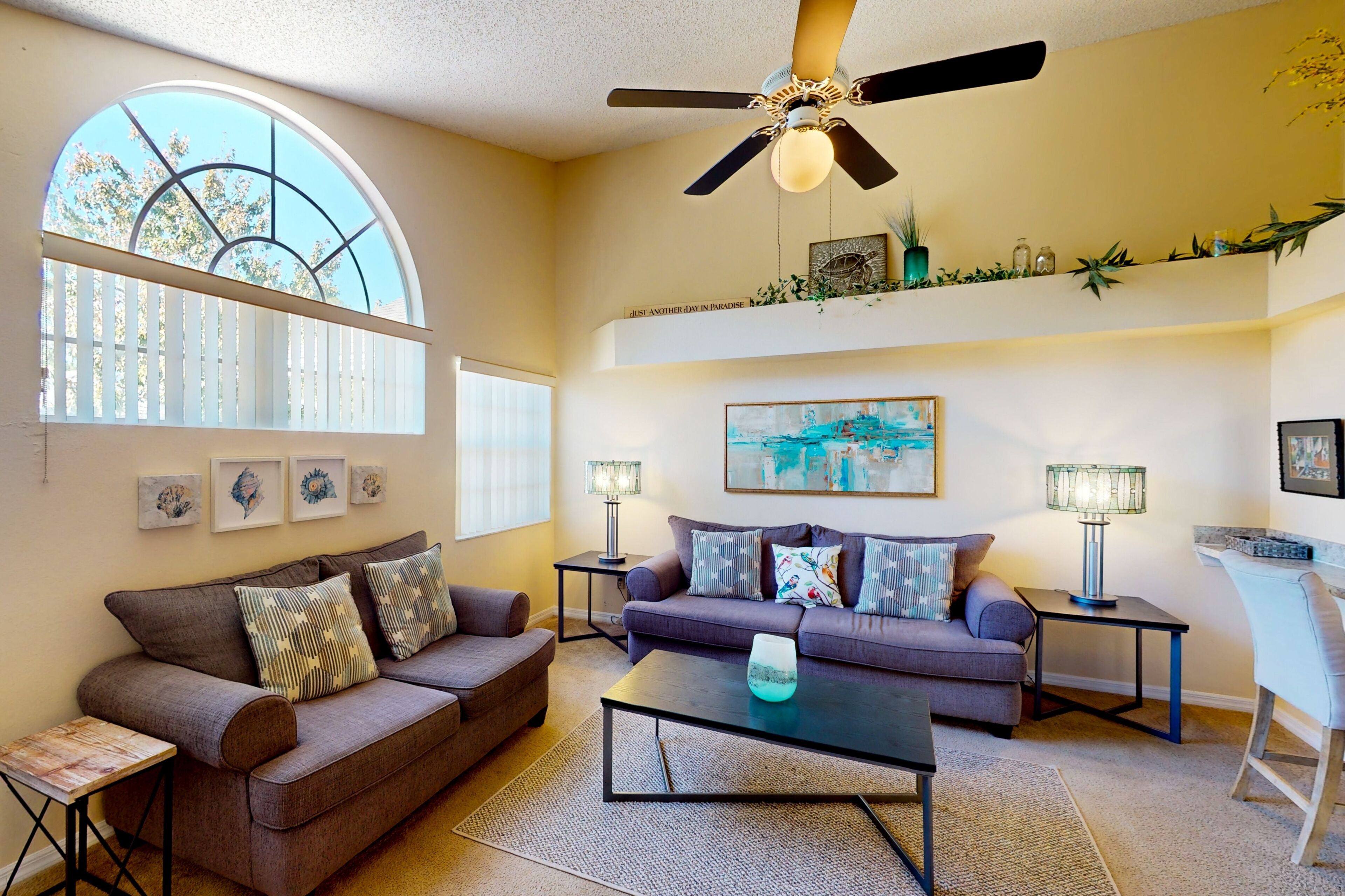 A living room with two sofas, a coffee table, a ceiling fan, and a large window with a view of trees.