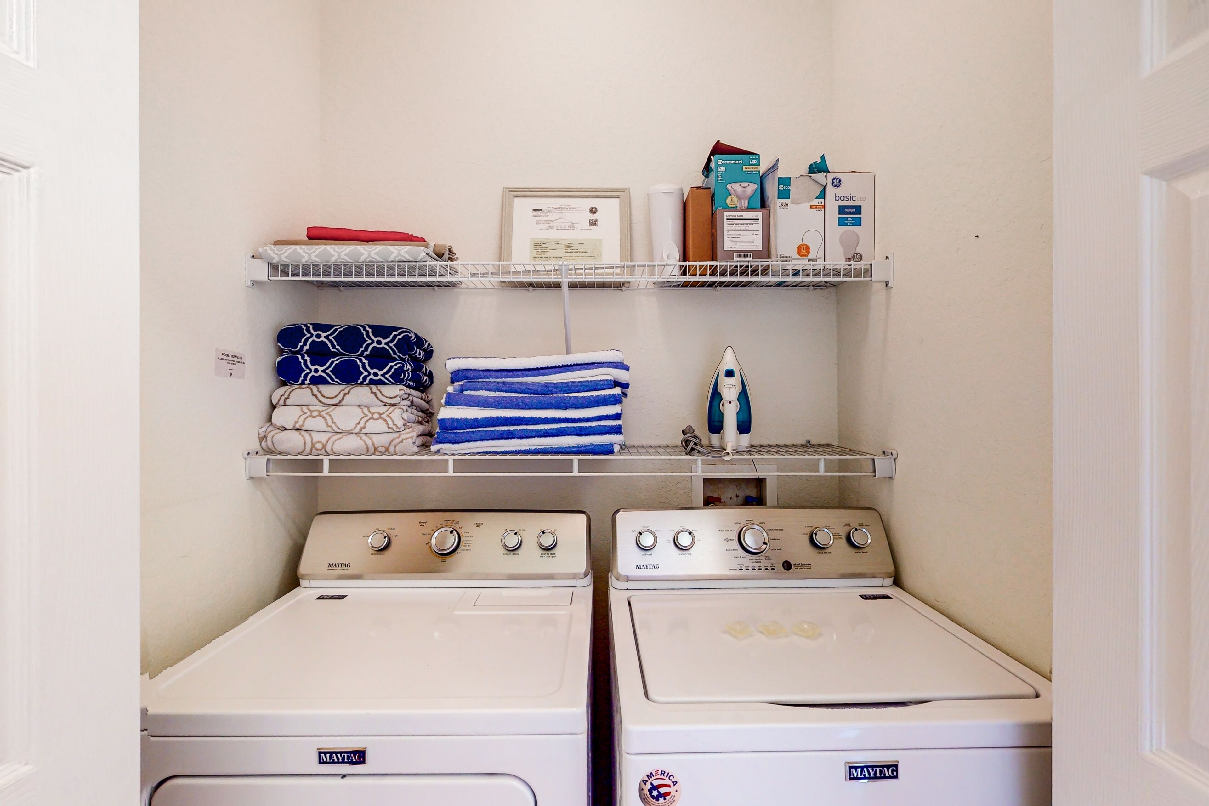A laundry area with two Maytag washing machines, a shelf with folded towels, a framed certificate, and laundry supplies.