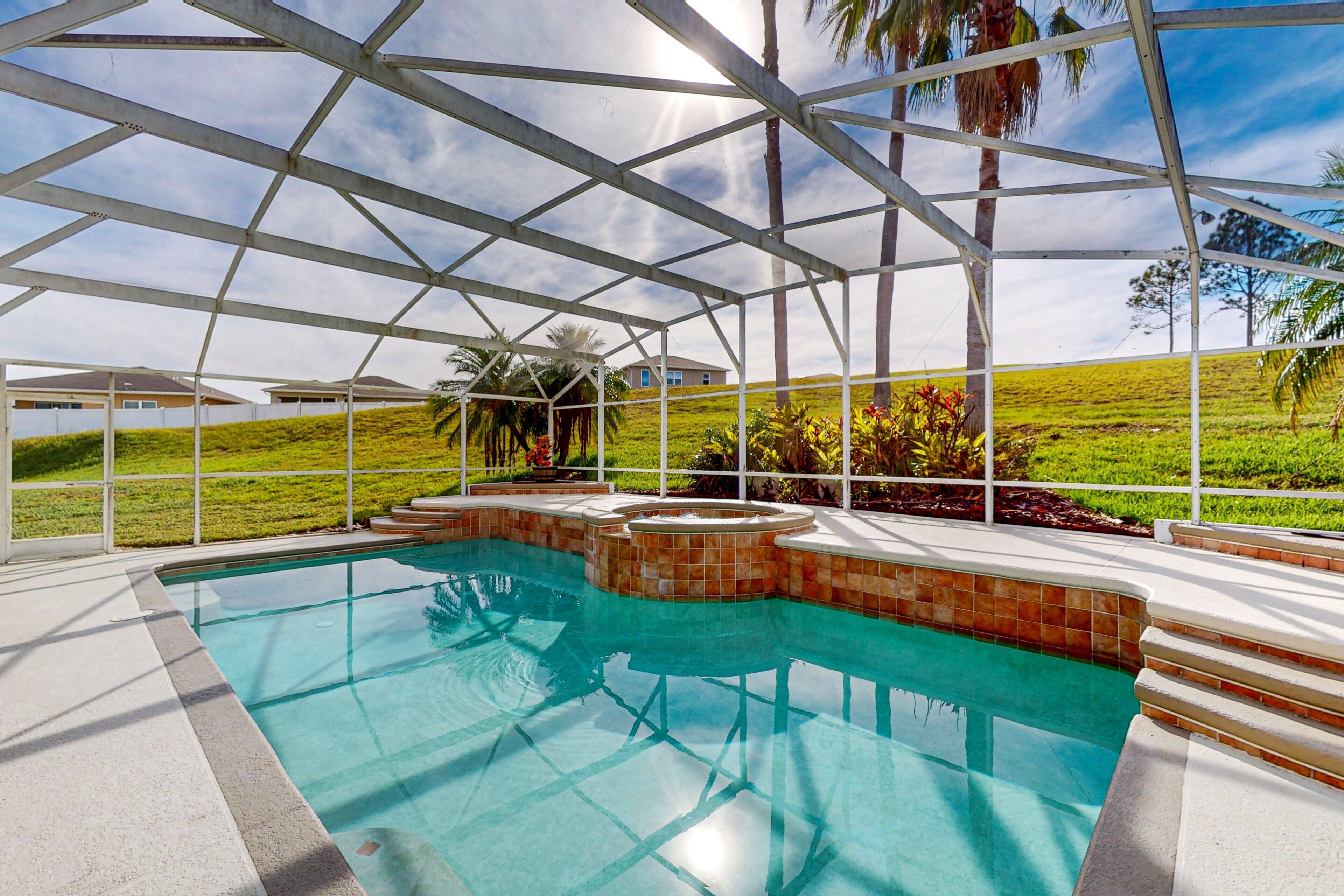 A swimming pool with a tiled edge, a covered area with a roof, and a view of a grassy landscape with palm trees.