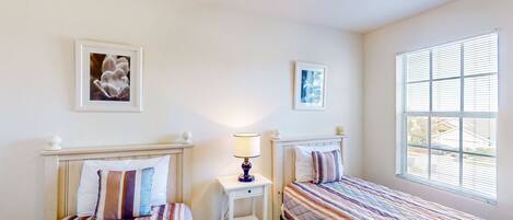 A bedroom with two beds, a nightstand with a lamp, and a window with blinds.