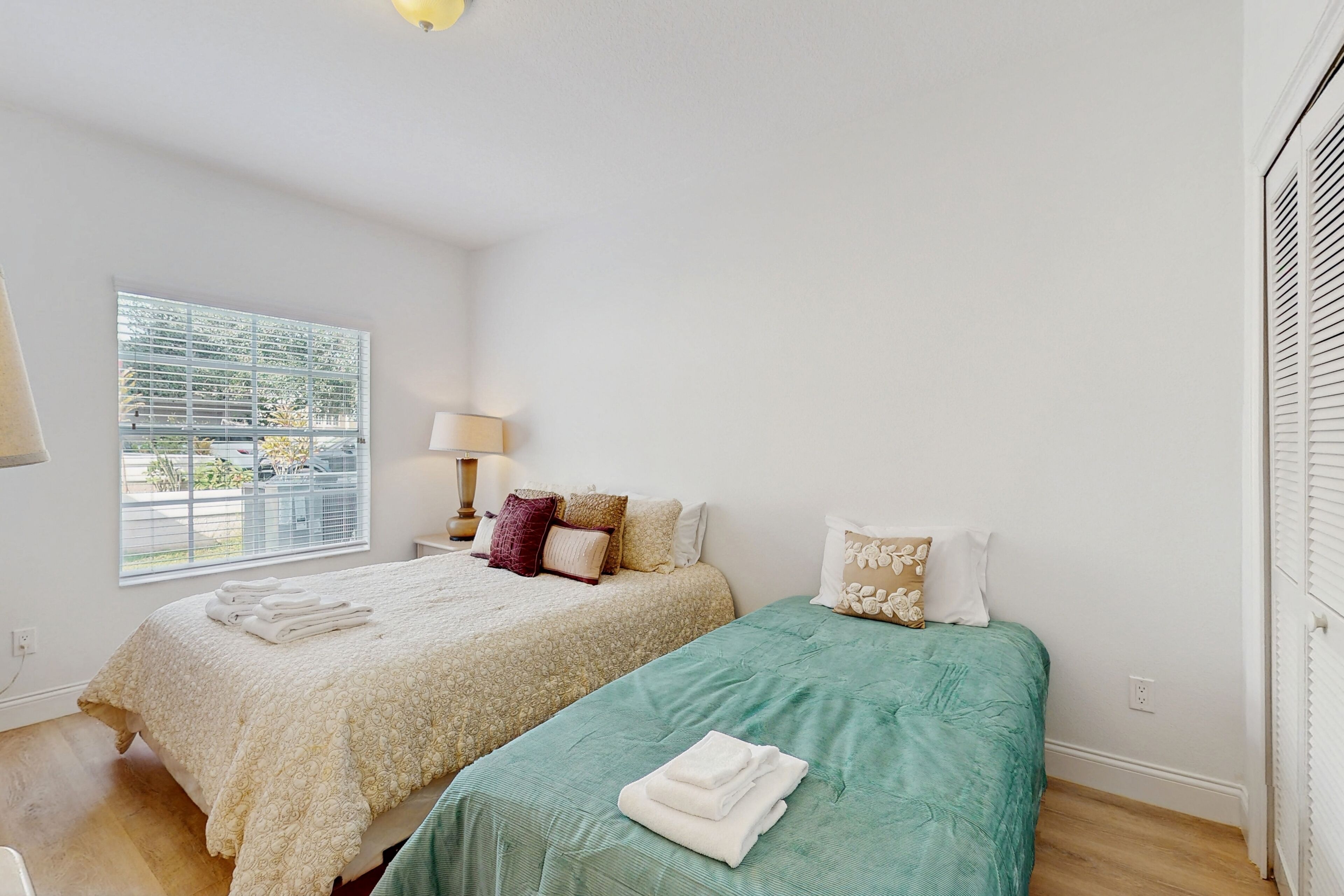 A bedroom with two beds, a window with blinds, a lamp, and a view of greenery outside.