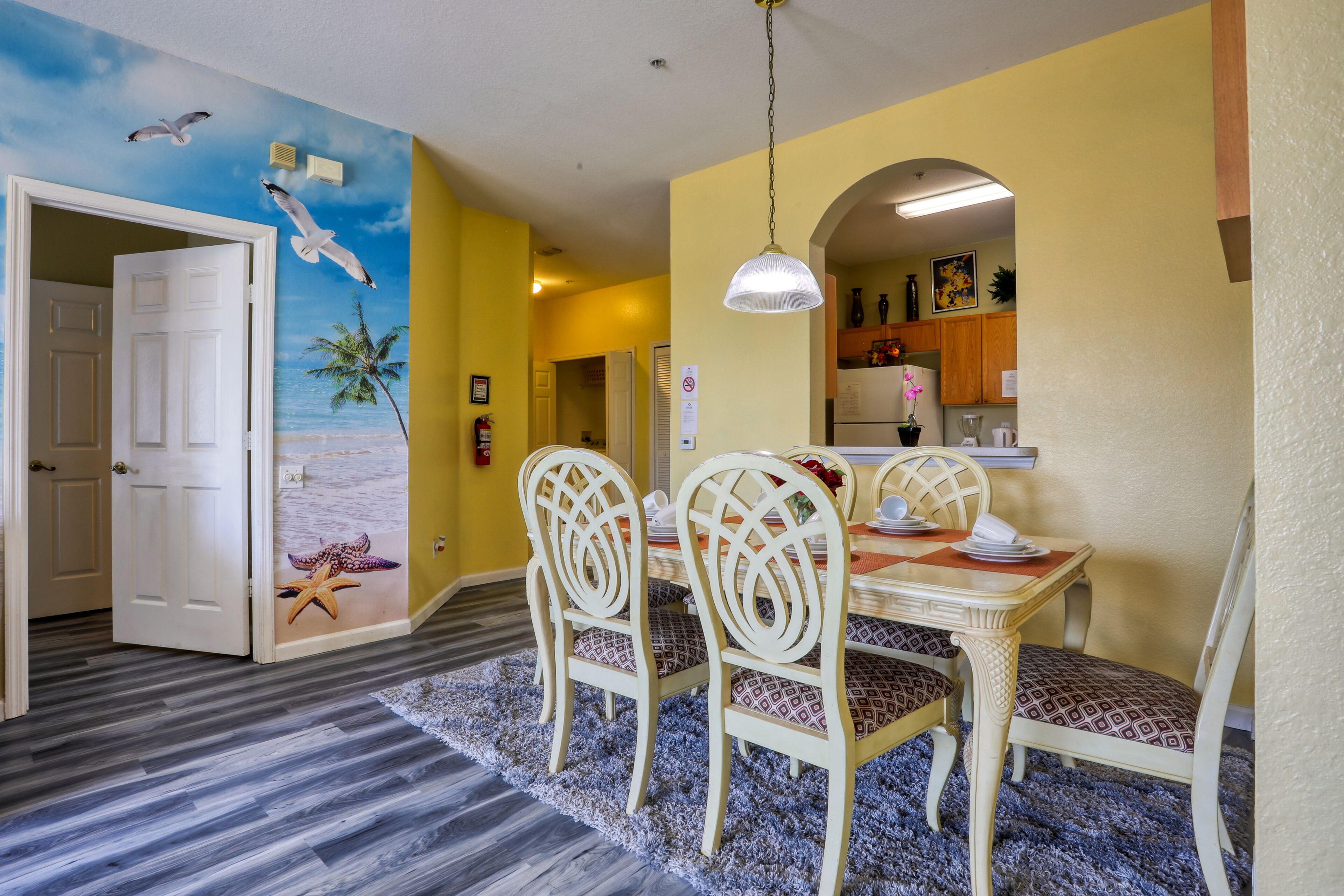 A dining area with a table set for four, white chairs, and a ceiling mural of seagulls and palm trees.