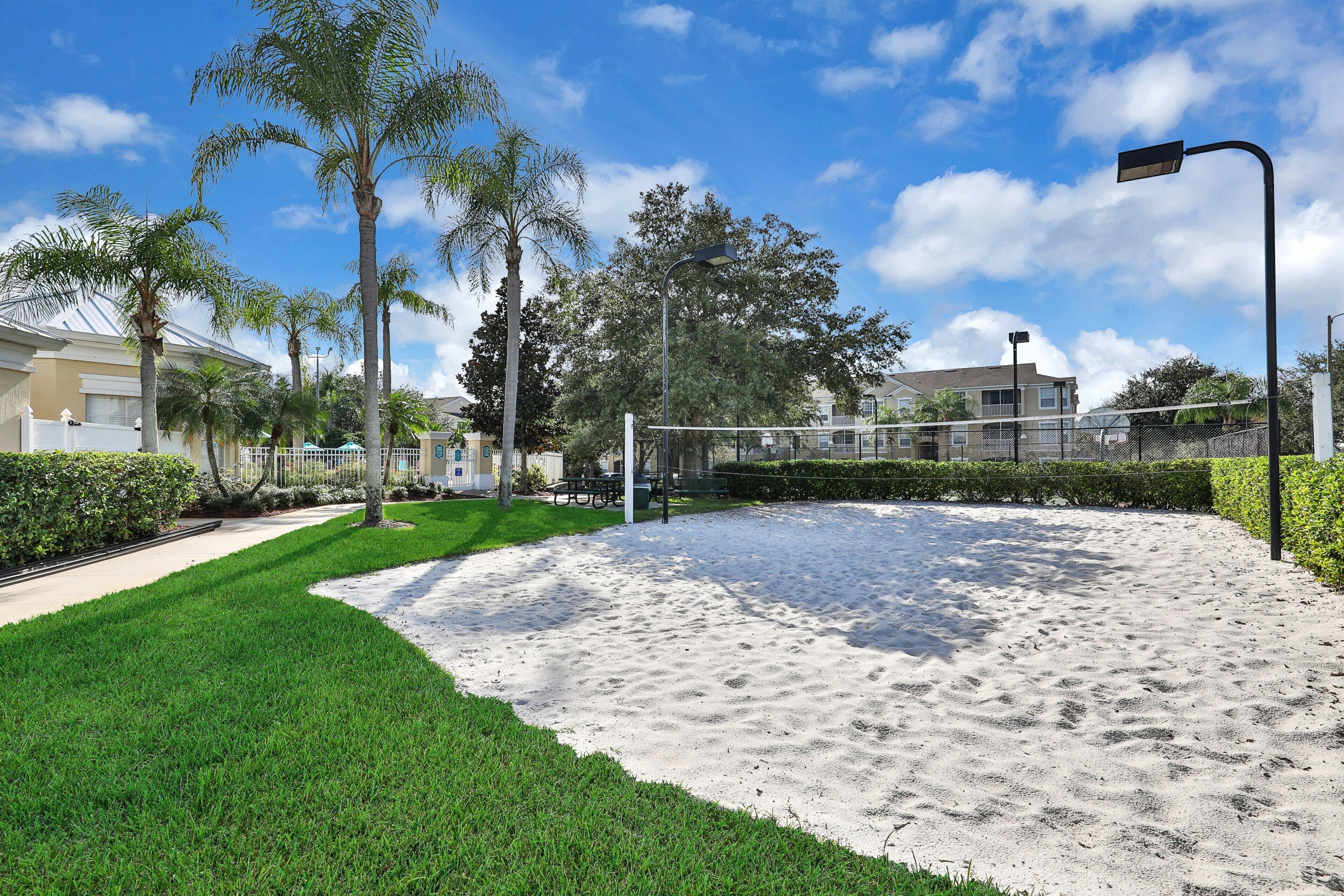 A sandy beach volleyball court with a net, surrounded by green grass and palm trees.