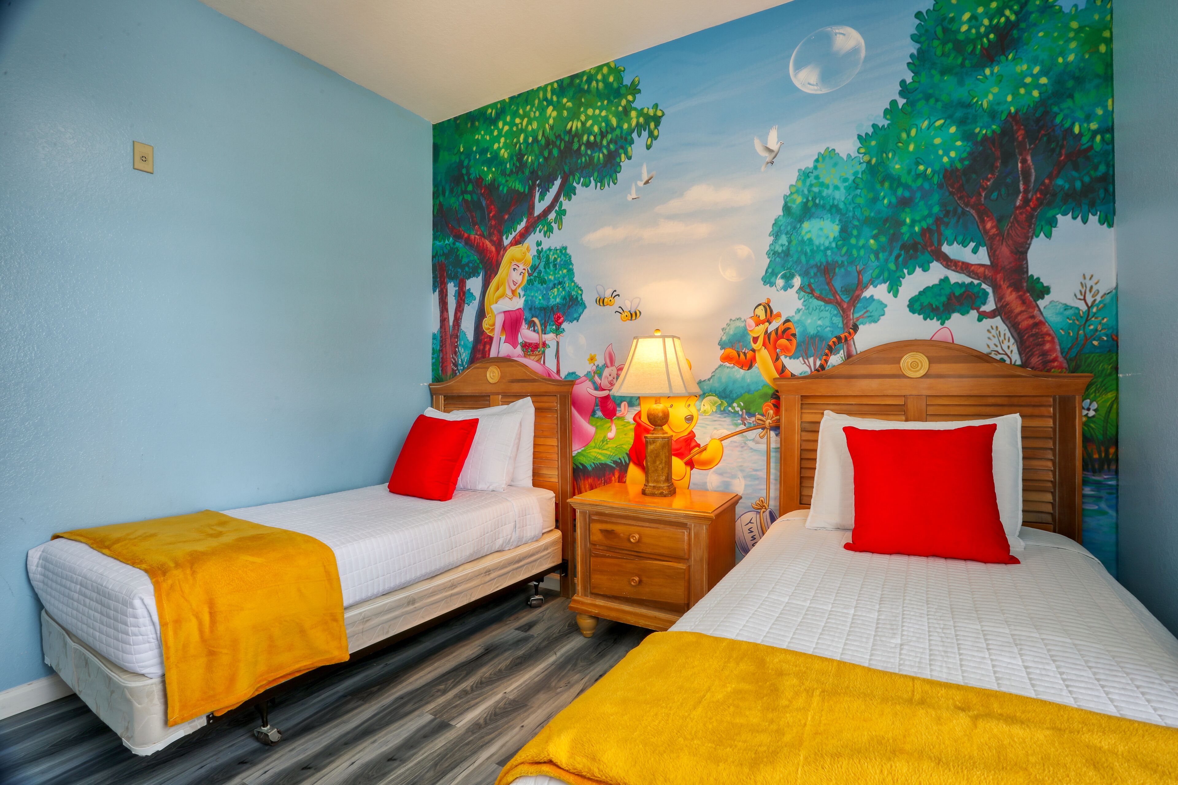 Ein Hotelzimmer mit zwei Betten, jeweils mit einer weißen Matratze und einer gelben Decke. Dazwischen steht ein Nachttisch aus Holz mit einer Lampe. An der Wand befindet sich ein Wandgemälde, das eine Waldszene mit Bäumen, Vögeln und einem Mond zeigt.