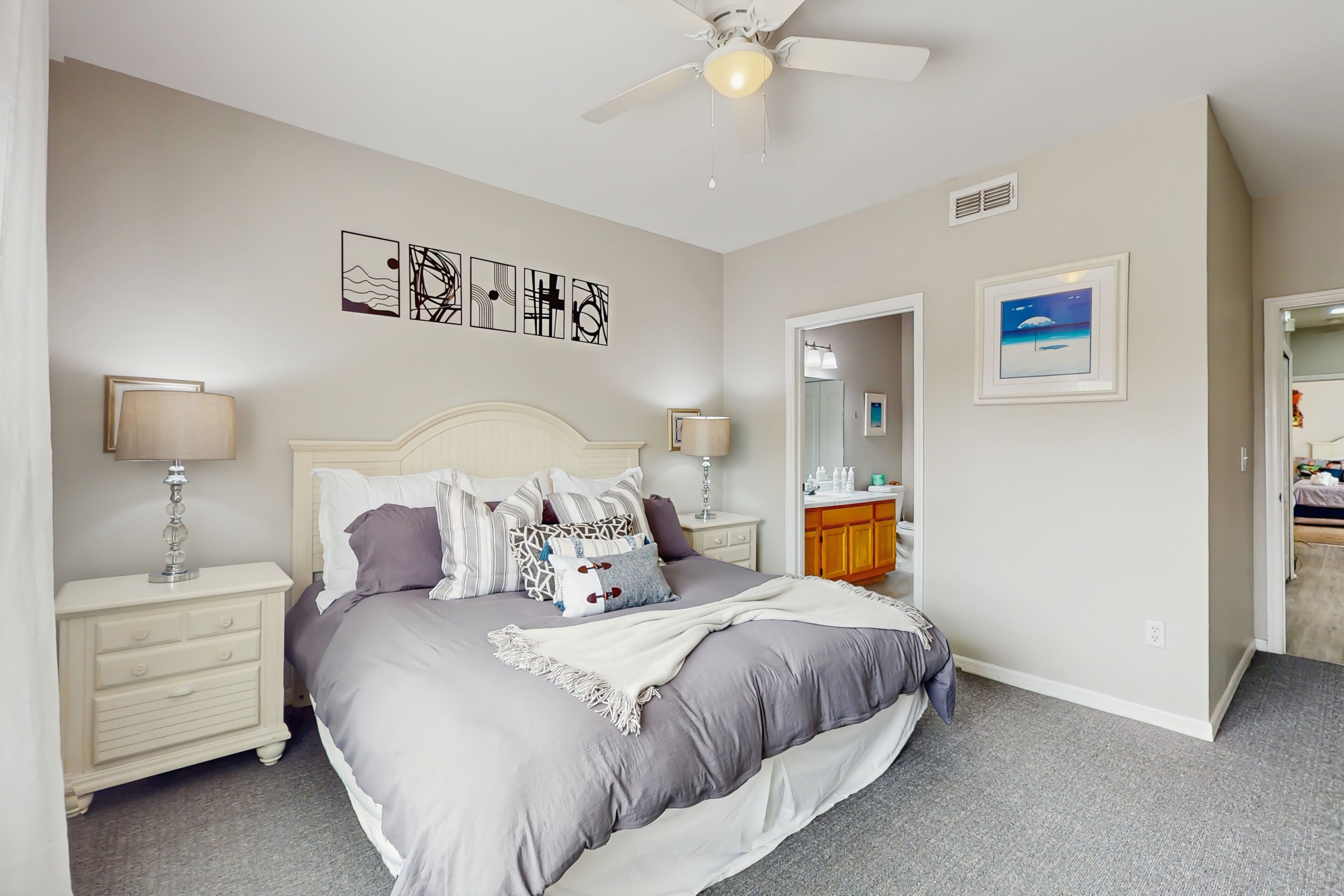 A bedroom with a bed, nightstands, a ceiling fan, artwork, and a bathroom visible through an open door.