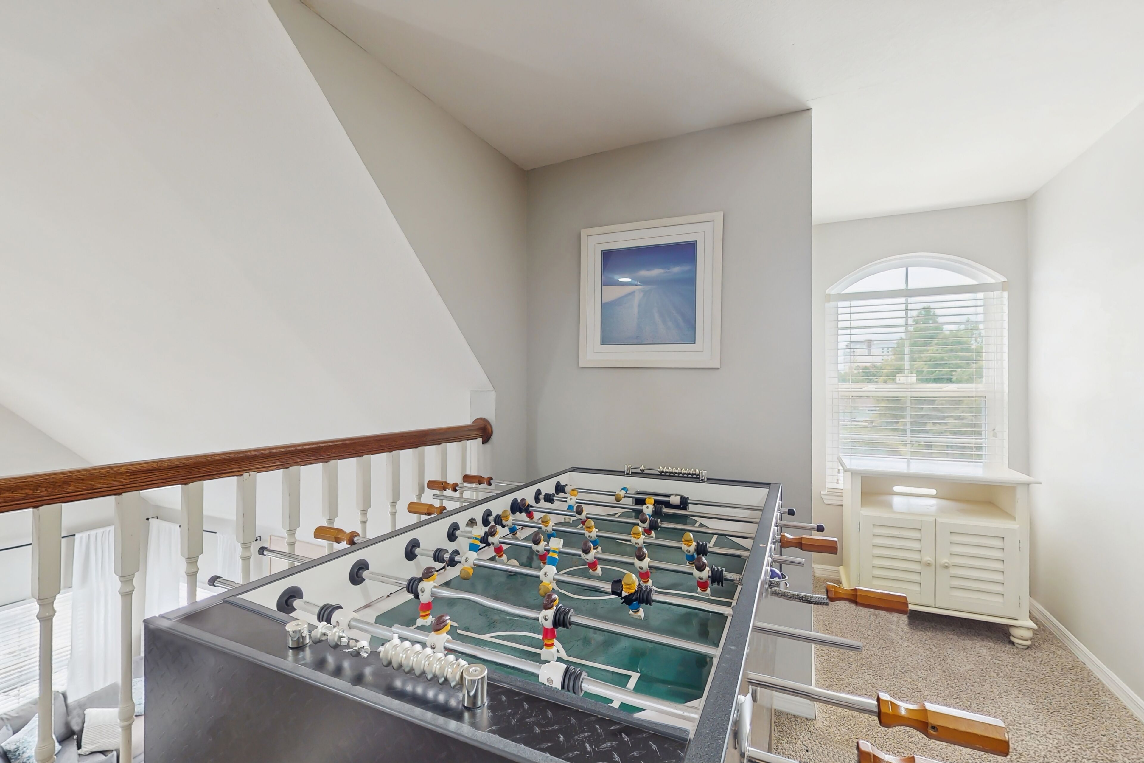 Una habitación amplia con mesa de fútbol, ventana con persianas y un pequeño armario blanco.