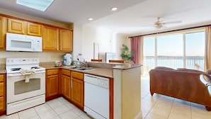 A kitchen with wooden cabinets, a microwave, an oven, and a dishwasher. There is a dining area with a brown sofa and a window with a view. - Blue Heron Beach I (Orlando)