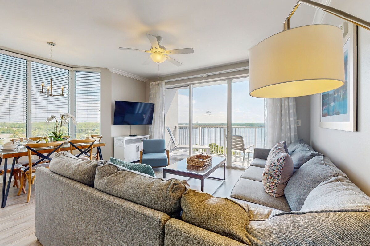 A spacious living room with a large sofa, a dining table, and a TV. There is a balcony with a view of the city.