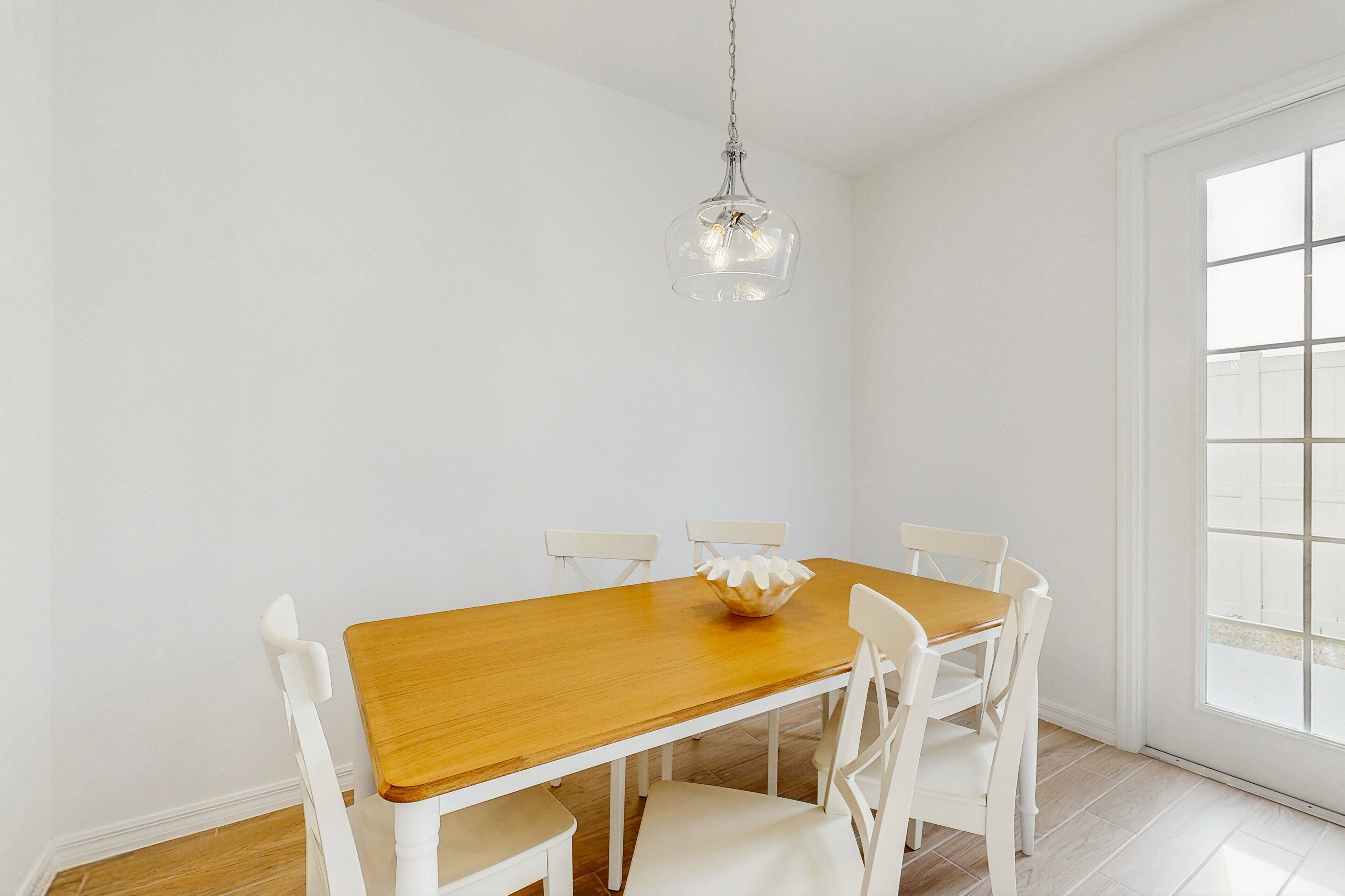 A dining area with a wooden table and white chairs, a hanging light fixture, and a large window.