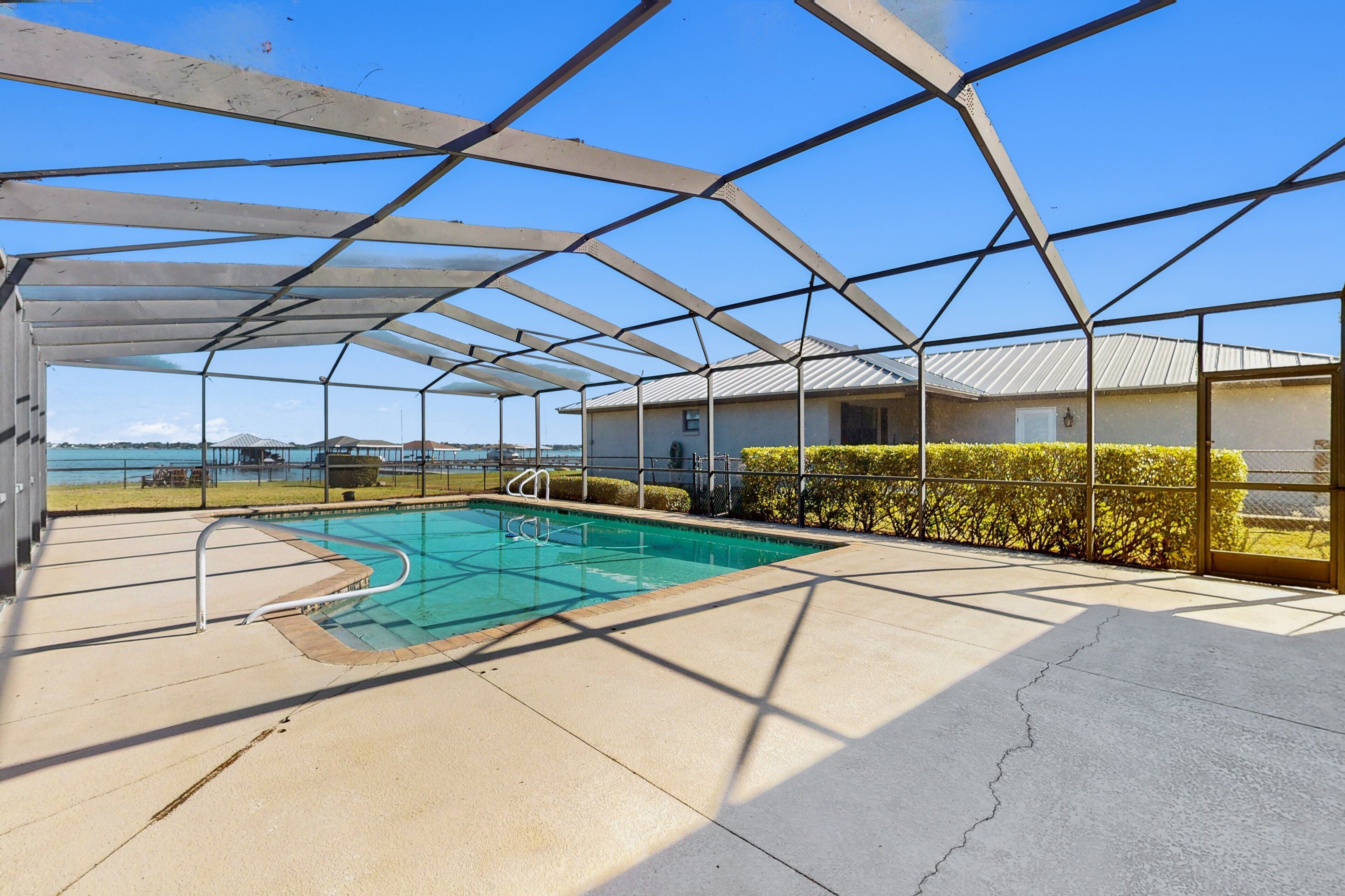 A covered outdoor swimming pool with a view of the sea, a building with a metal roof, and a hedge.