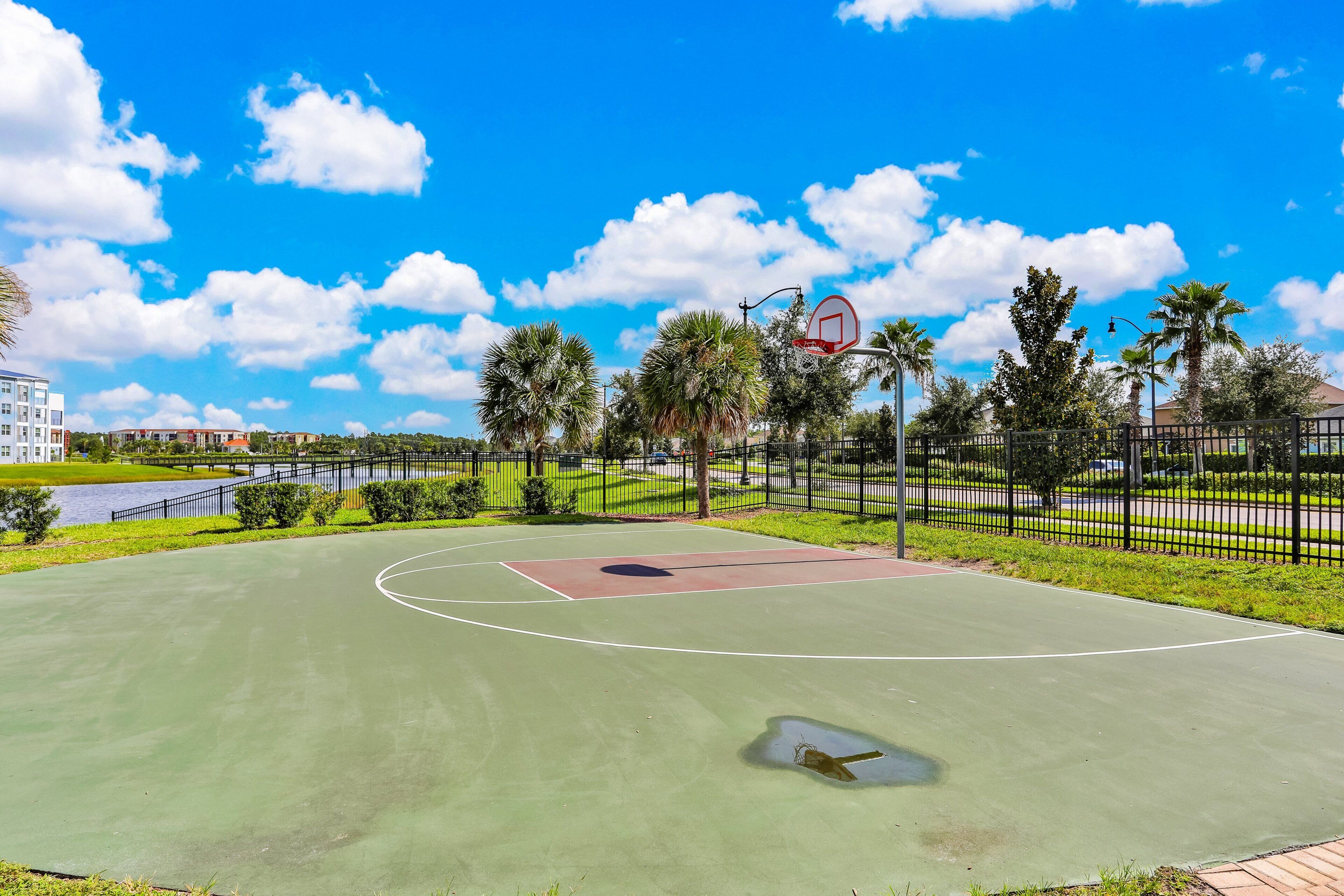 Ein Basketballplatz im Freien mit blauem Himmel, umgeben von Bäumen und einem Zaun.