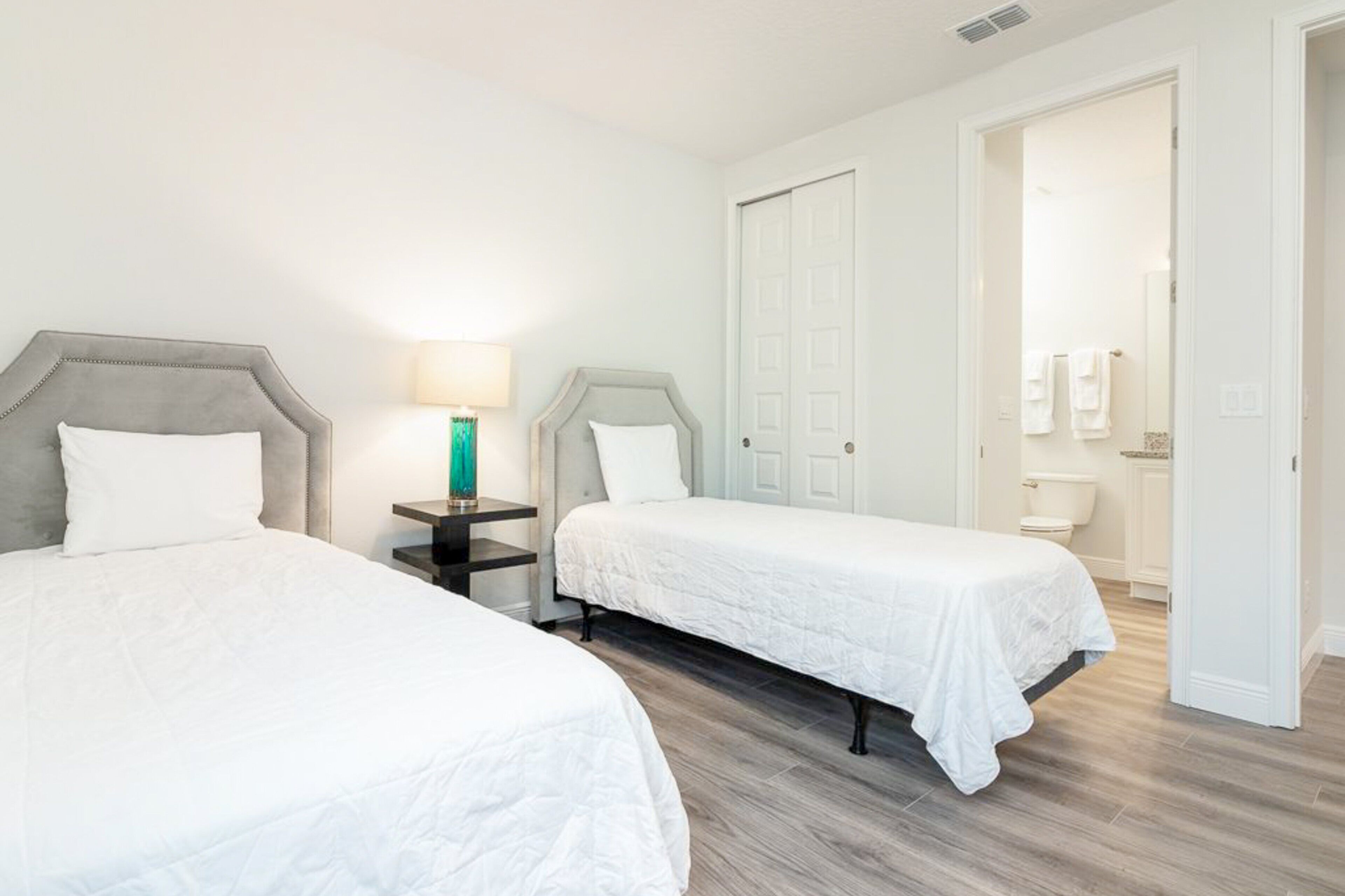 Habitación de hotel con dos camas, una mesita de noche con lámpara, una puerta que da a un baño y un inodoro a la vista.