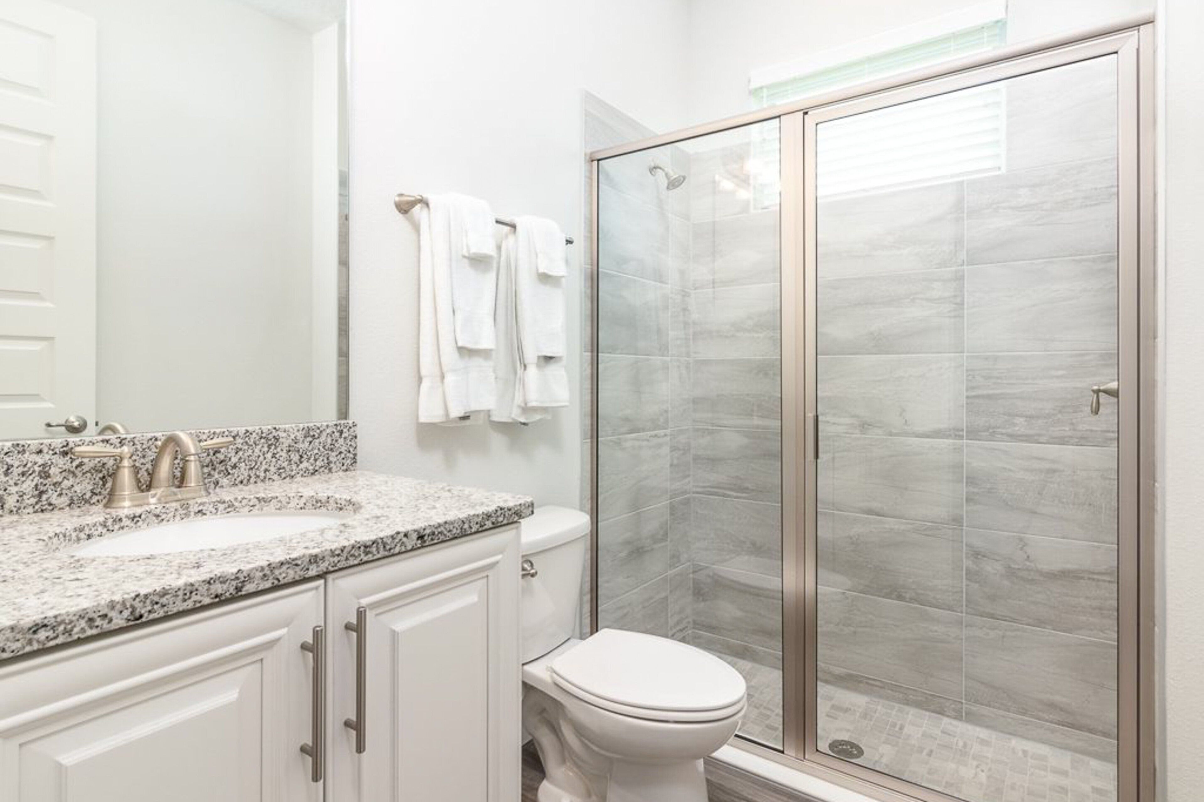 Une salle de bain avec un lavabo sur un plan de travail en granit, des meubles blancs, des toilettes et une douche à parois de verre.