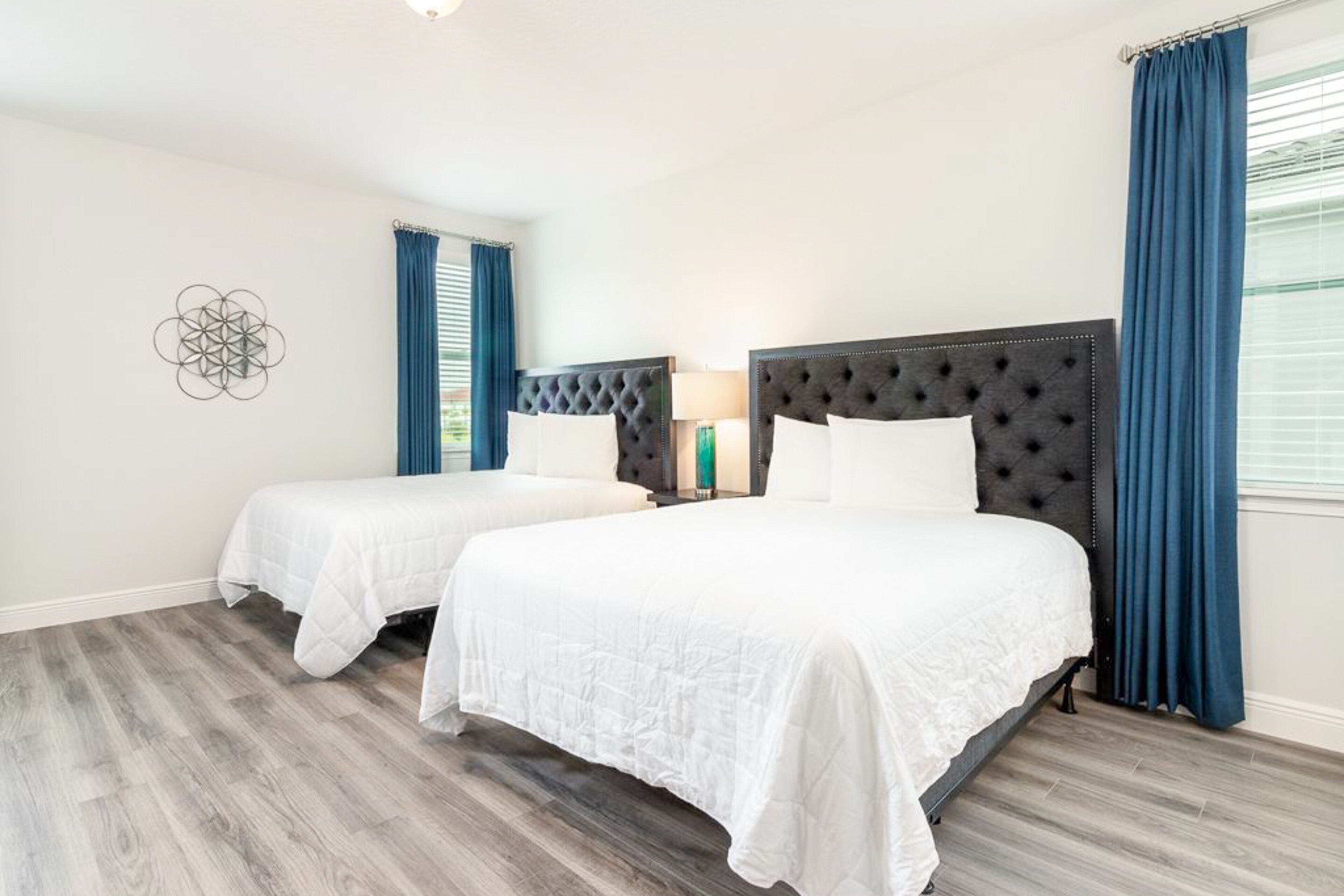 A hotel room with two beds, a decorative wall piece, a nightstand with a lamp, and blue curtains.