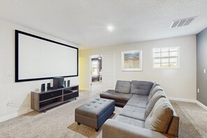 A living room with a large flat-screen TV, a gray sectional sofa, a small ottoman, and a wooden entertainment center. - Modern Home w/ Private Pool & Spa, Theater, Game Room, and Resort Amenities (Davenport)