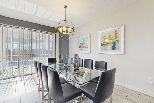 A dining area with a glass table, black chairs, a chandelier, and two framed artworks on the wall. - Modern Home w/ Private Pool & Spa, Theater, Game Room, and Resort Amenities (Davenport)