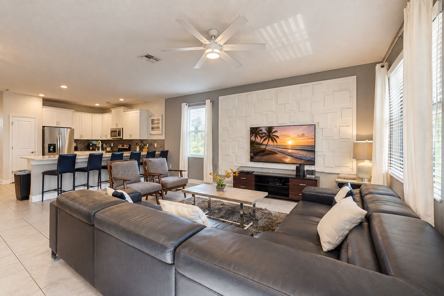 A modern living room with a sectional sofa, a flat-screen TV mounted on the wall, and a kitchen in the background.