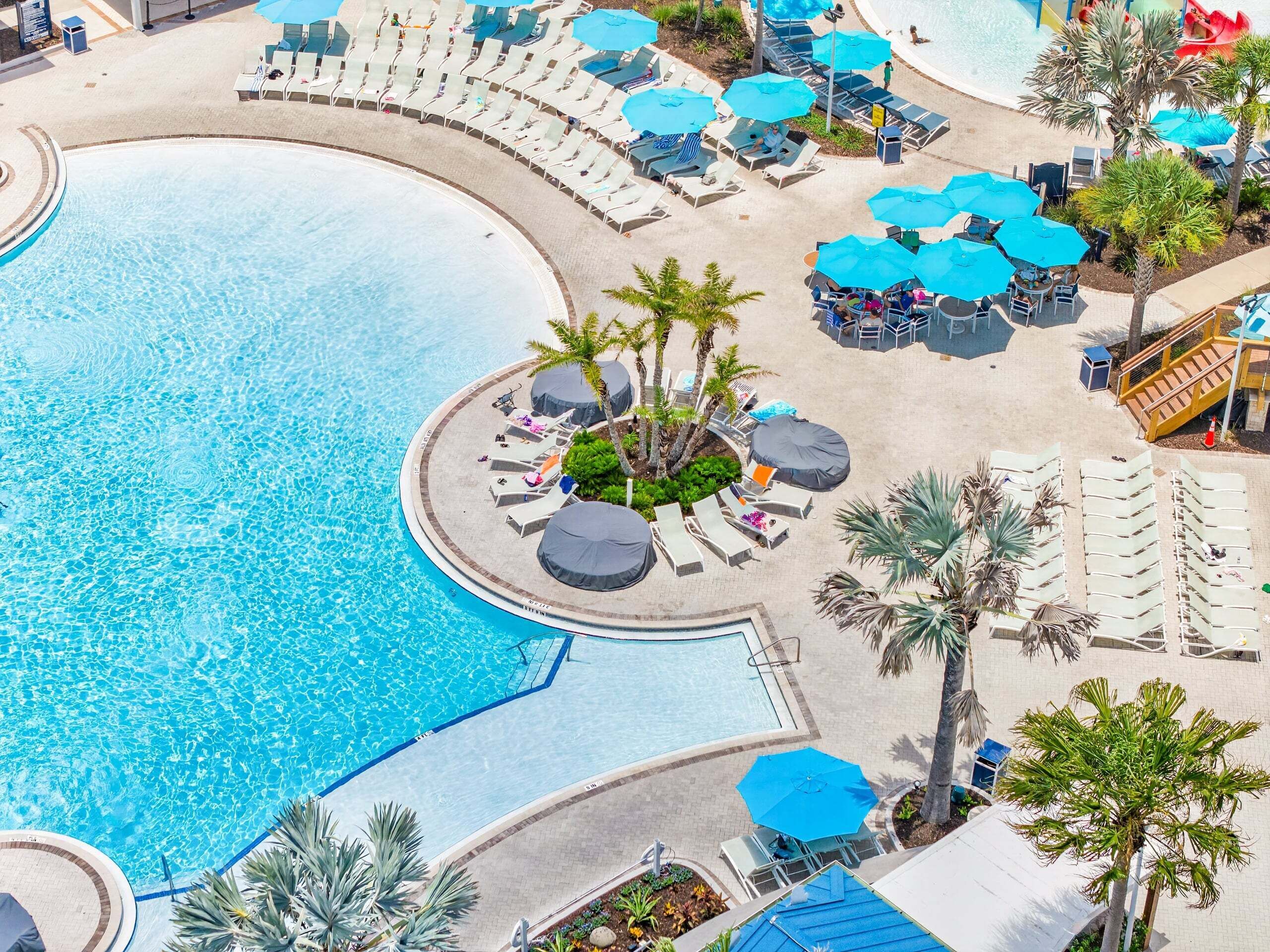 A large pool with sun loungers and umbrellas, surrounded by palm trees and a small waterfall feature.